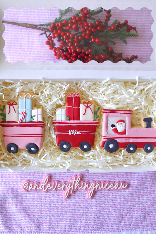 Christmas train-themed three-part pink and red cookie in a white gift box with clear window, styled on pink and white striped fabric, hand-decorated, perfect holiday gift.