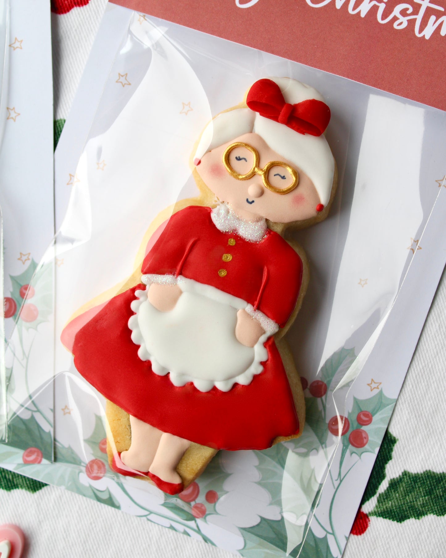 Close-up of a Mrs Claus Christmas cookie wearing a tea-length red dress with a white collar and gold buttons, her white hair styled in a bun with a red bow.