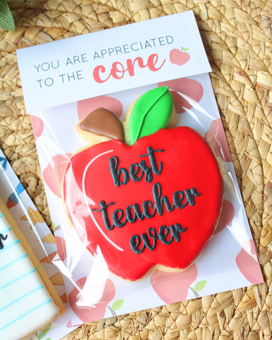 Close-up of a teacher appreciation cookie styled on a natural hyacinth placemat with eucalyptus sprigs. The cookie is shaped like a red apple with a green leaf and the words “best teacher ever” in black script icing. The apple cookie’s topper reads “You are appreciated to the core.