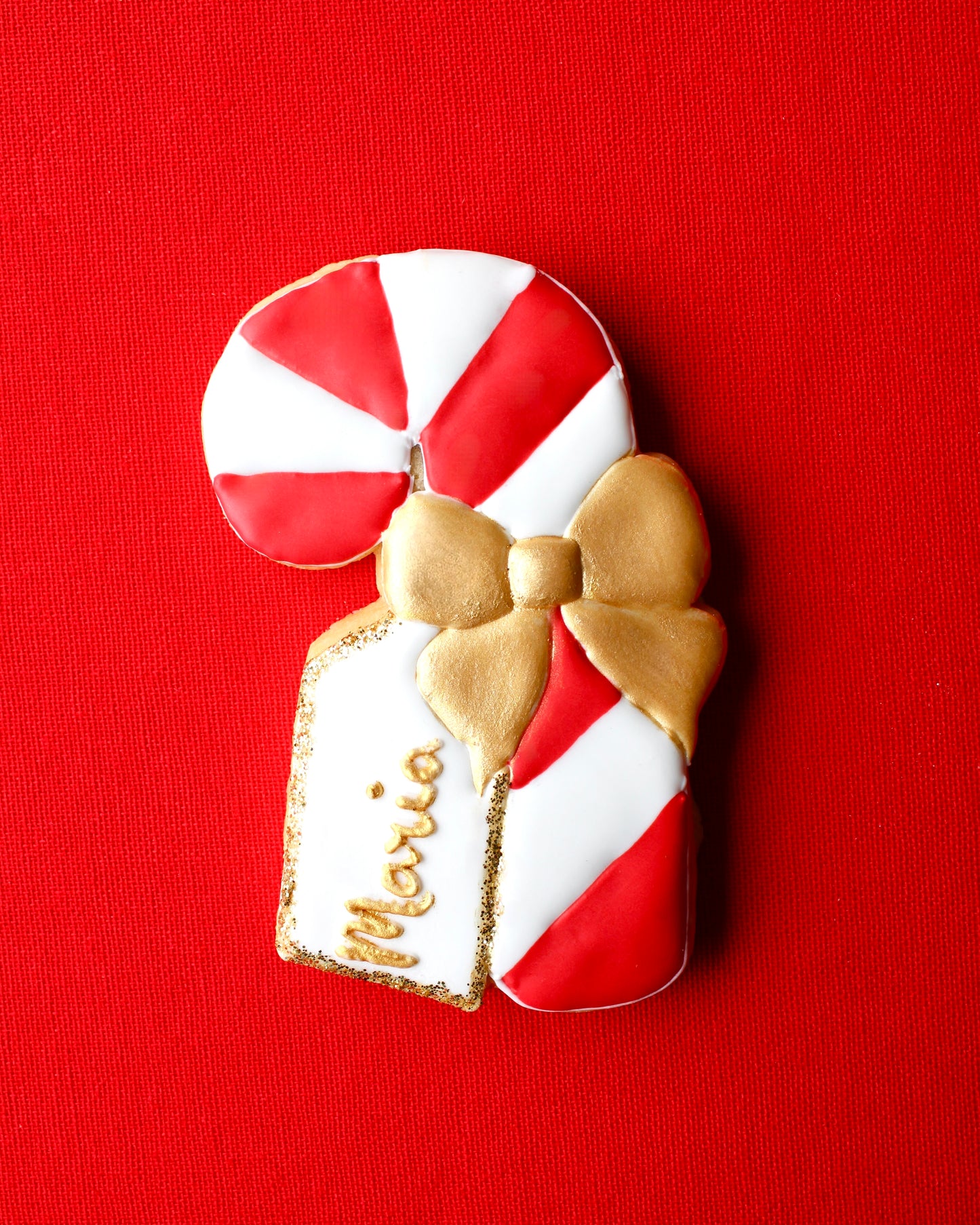 Close-up of a red and white candy cane cookie decorated with a gold icing bow and a white edible name tag that says "Maria" in gold lettering with edible gold glitter edges, styled on red linen fabric.