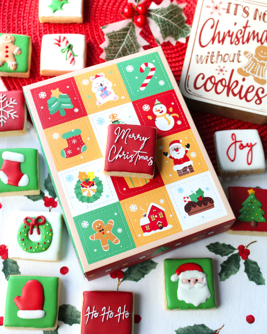 12 Days of Christmas cookie box featuring 12 mini Christmas-themed cookies arranged around the box, with a red cookie on top reading “Merry Christmas” in white cursive lettering. The cookies are decorated in festive red, green, white, and gold colours.