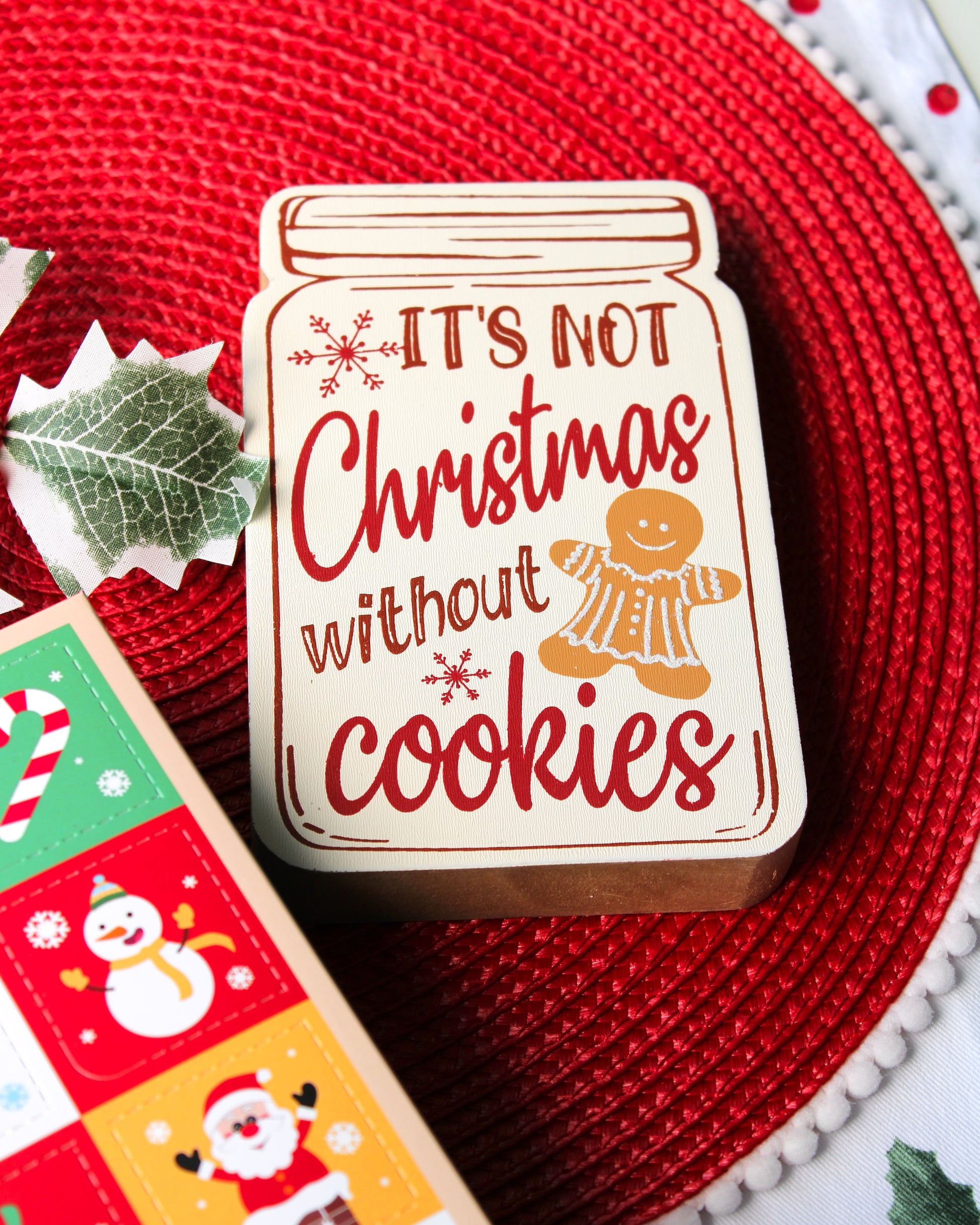 Wooden sign shaped like a mason jar that reads “It’s not Christmas without cookies,” featuring a gingerbread cookie person design, styled on a red placemat.