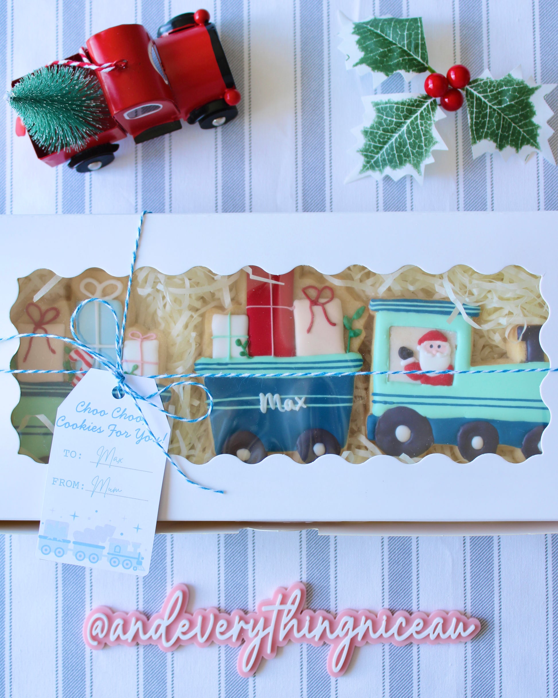 Christmas train-themed three-part blue and mint green cookie in a white gift box with clear window, hand-decorated with red festive details, styled on grey and white striped fabric, perfect holiday gift.