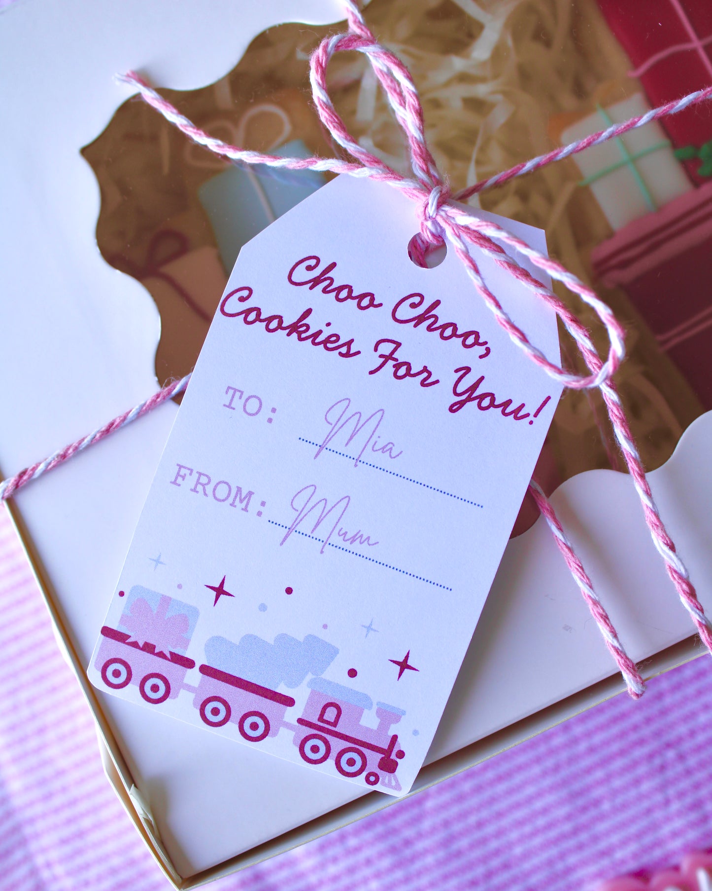 Close-up of a gift tag tied to a cookie box with pink and white twine, reading “Choo choo, cookies for you! To: Mia From: Mum,” festive holiday packaging.