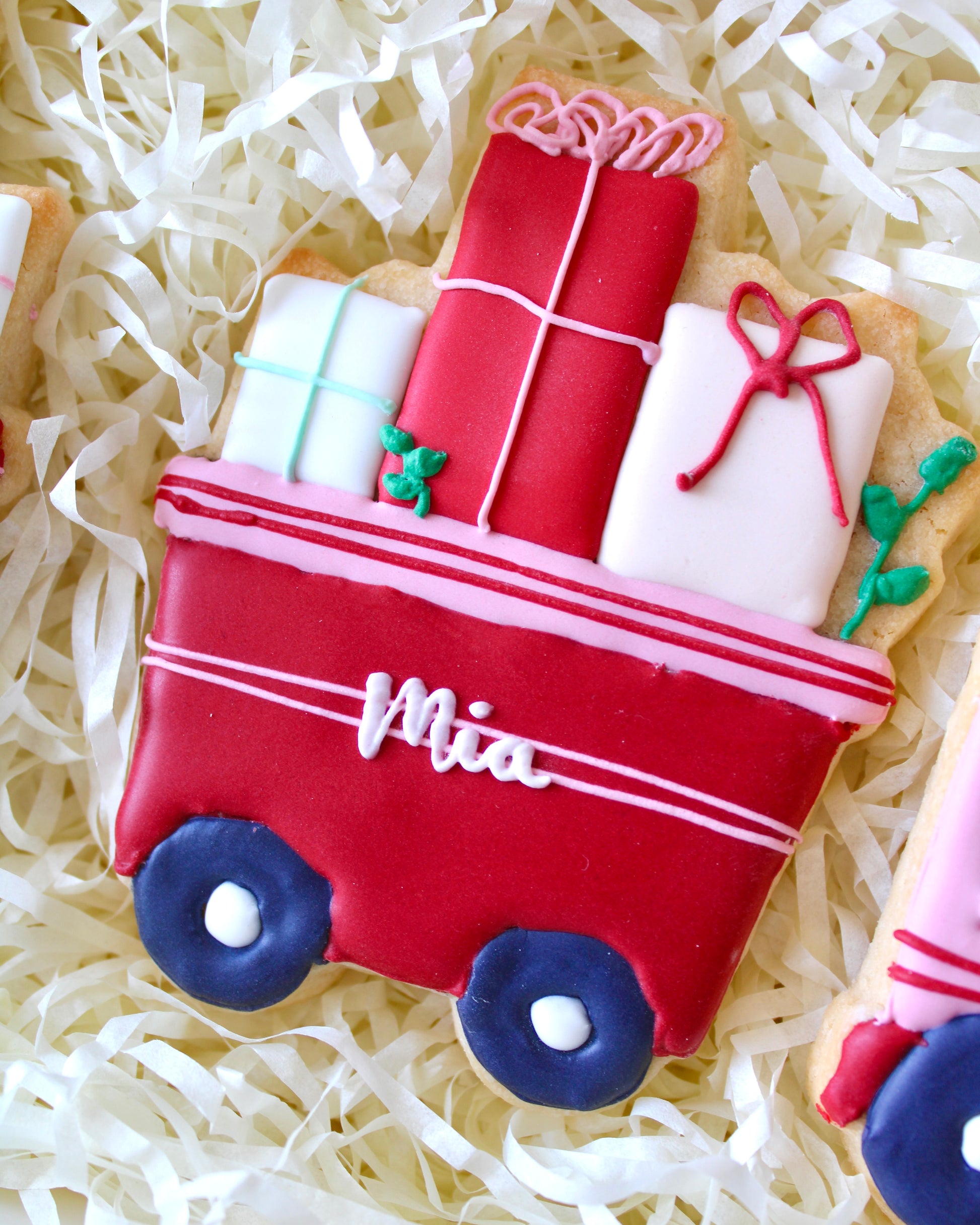 Close-up of a red train cart cookie with the name "Mia" piped in white icing, filled with royal icing presents, placed on white shredded paper in a white gift box, festive holiday treat.