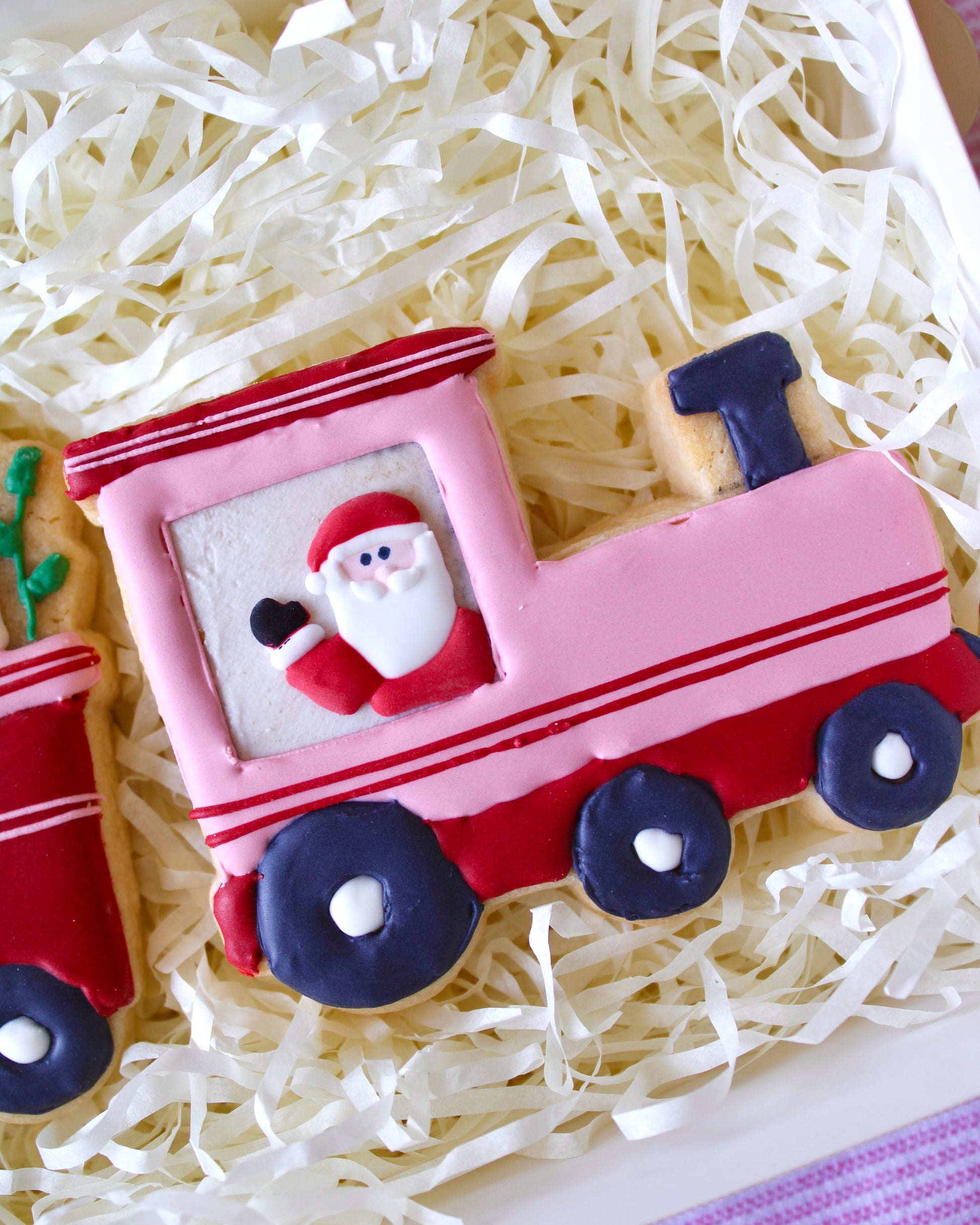 Close-up of a pink train cookie with red accents, featuring Santa waving from the driver’s window, placed on white shredded paper in a white gift box, festive holiday treat.