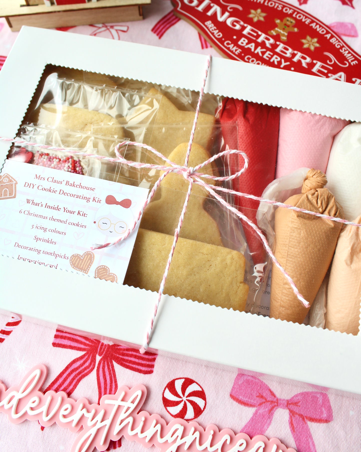 White gift box filled with a DIY Christmas cookie decorating kit, tied with pink and white twine. Inside are individually wrapped cookies, piping bags filled with royal icing, and a packet of festive sprinkles.