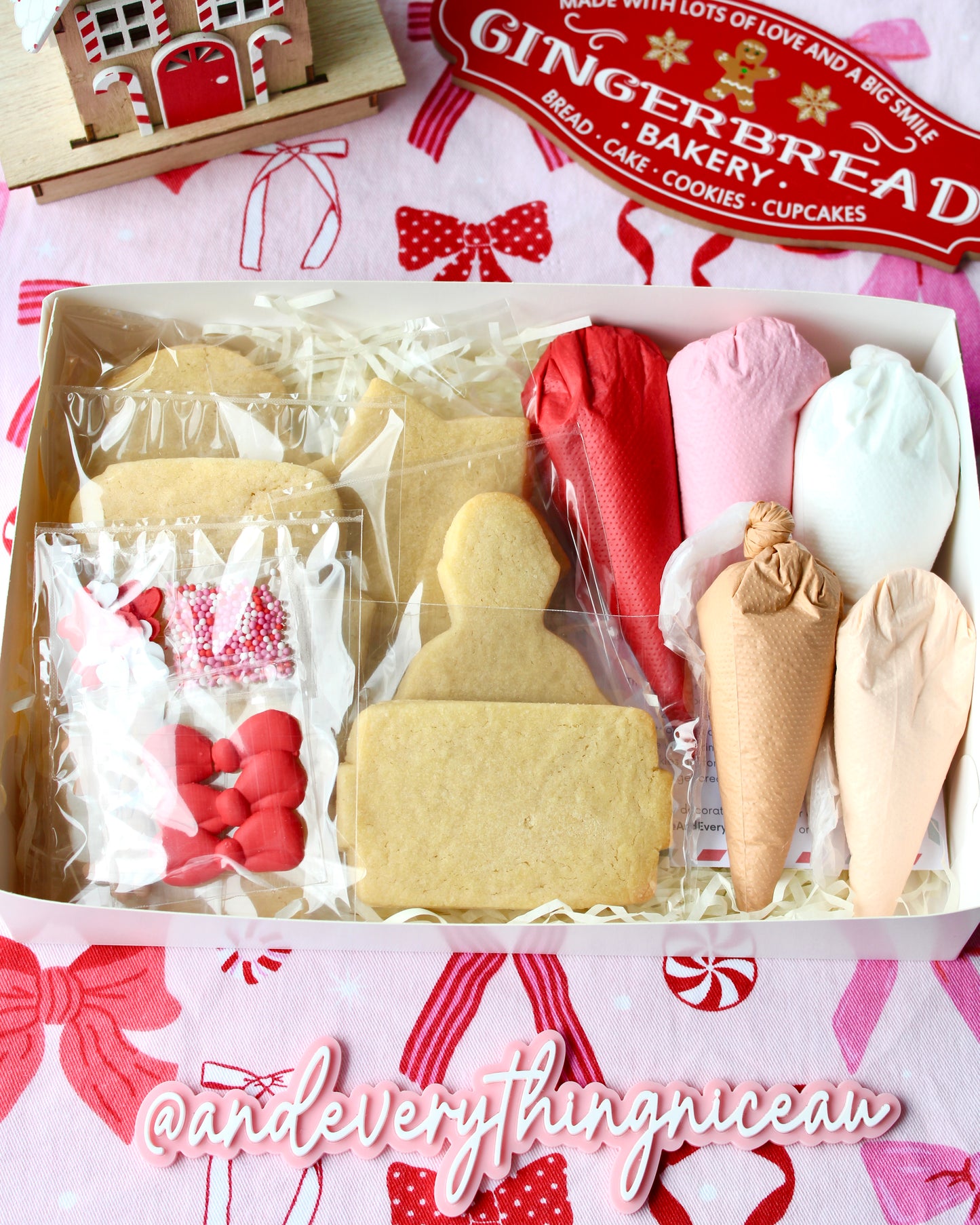 DIY Christmas cookie decorating kit laid out on pink and red bow-patterned fabric, featuring six individually wrapped vanilla cookies in festive shapes, four piping bags of royal icing in red, pink, white, tan, and cream, assorted sprinkles, a decorating pick, and an instruction card.