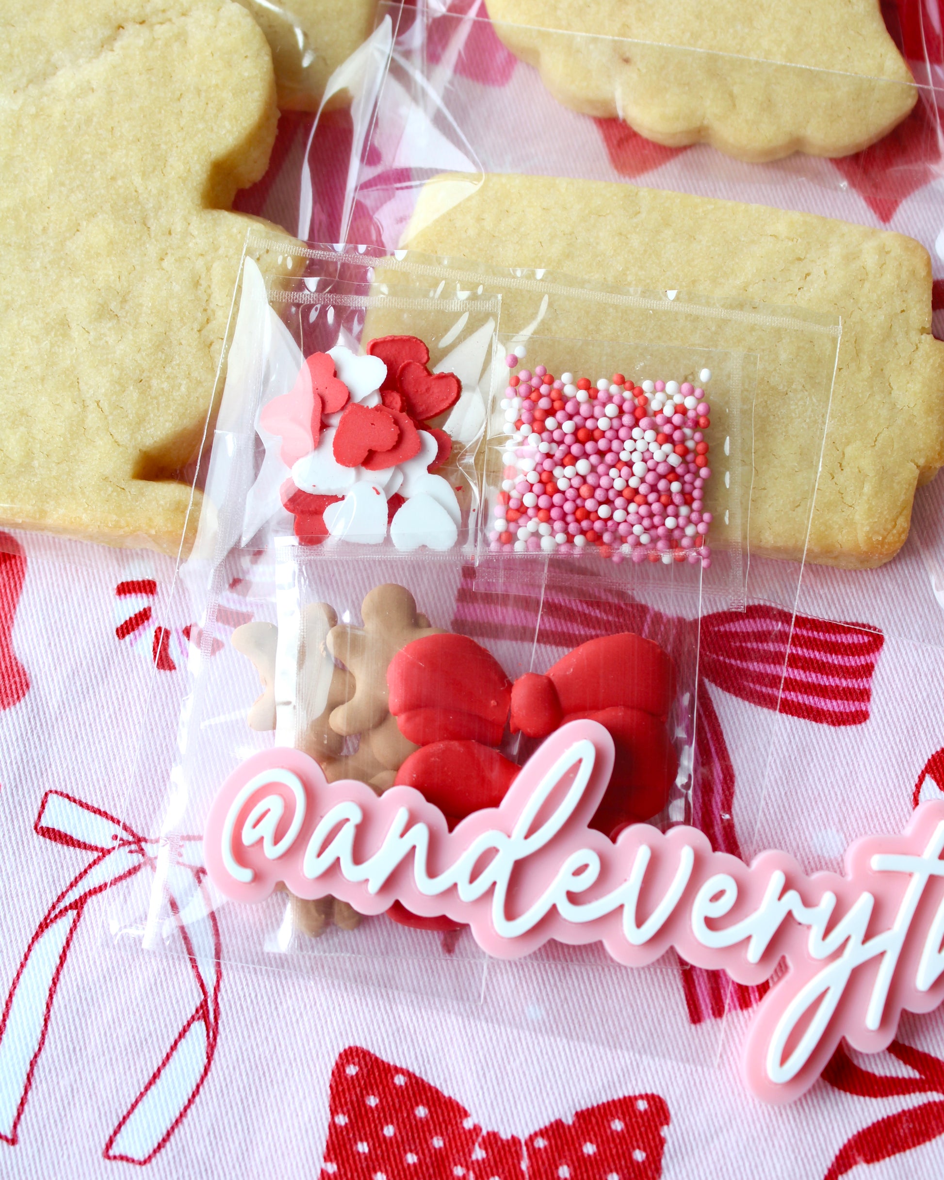 Close-up of a sprinkle packet filled with assorted pink, red, and white sprinkles, displayed on pink and red bow-patterned fabric with two cookies visible in the background.