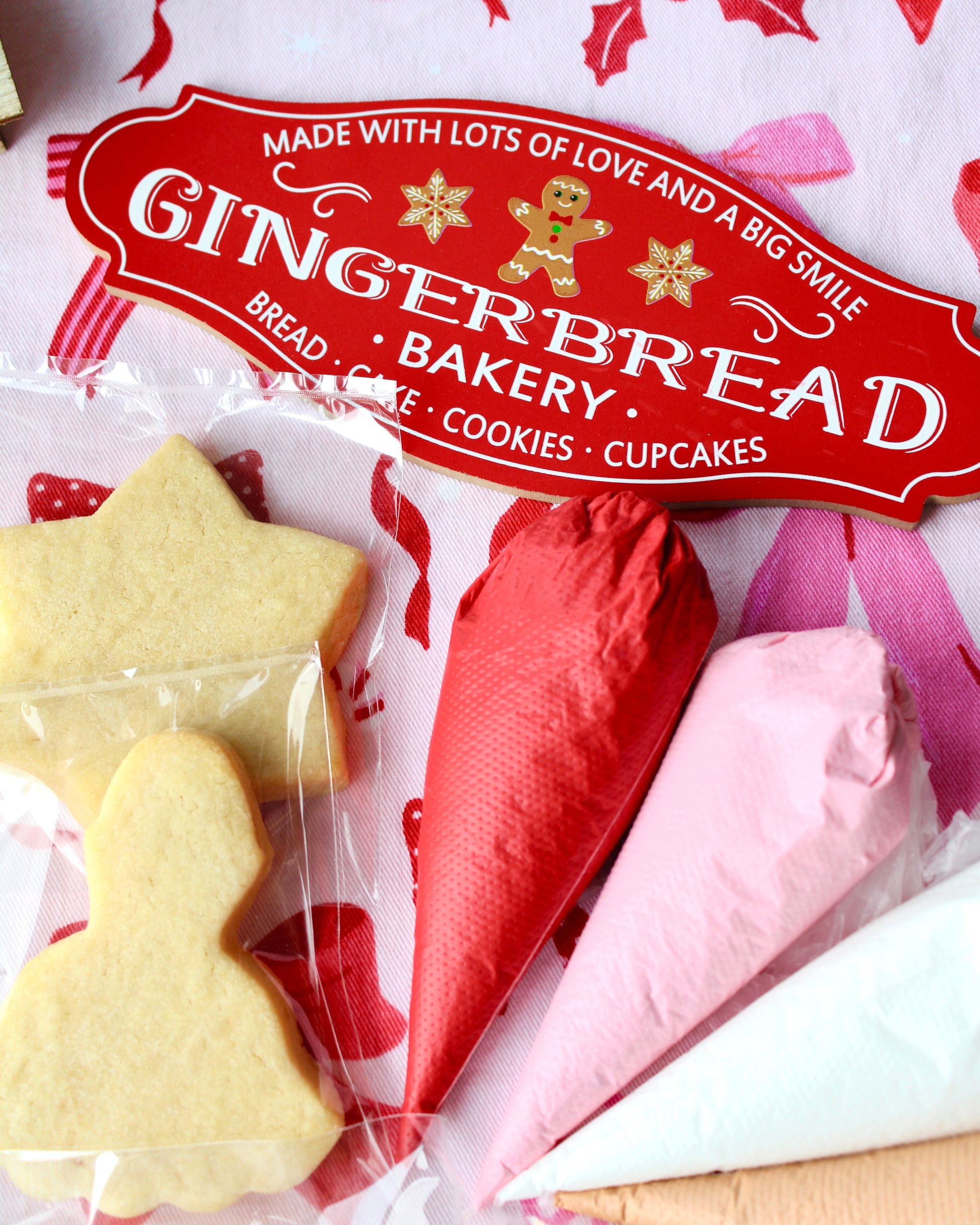 Close-up of a red Christmas sign reading “Gingerbread Bakery,” with red and pink icing bags visible in the corner and two cookies in view, set on pink and red bow-patterned fabric.