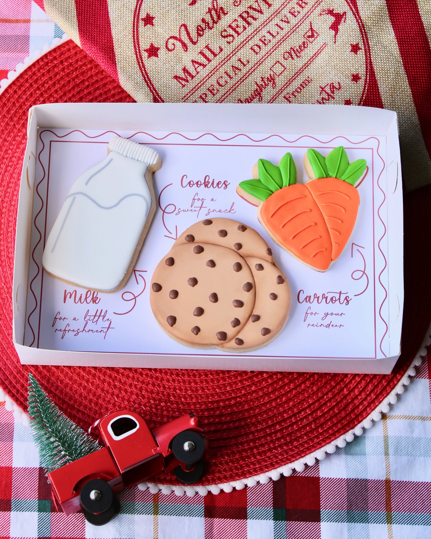 Three large Christmas cookies from the Santa’s Treat Box displayed in a white gift box. The cookies include a bottle of milk, a stack of cookies, and a bunch of carrots. The box sits on a red placemat over red and green tartan fabric, with a vintage mail-themed stocking in the top right corner.