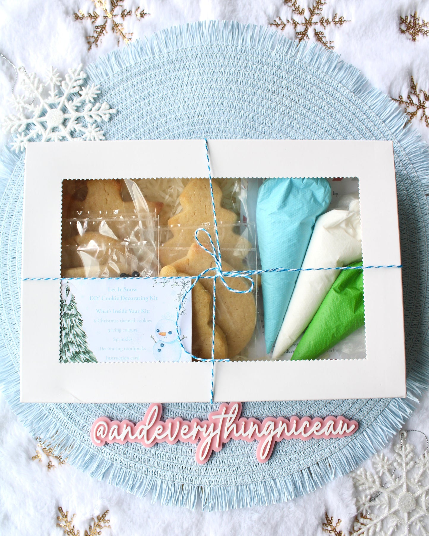 Christmas DIY cookie decorating kit in a white gift box tied with blue and white twine, displayed on a pale blue placemat over fluffy white fabric with gold snowflakes. Winter-themed styling with a festive, cozy feel.