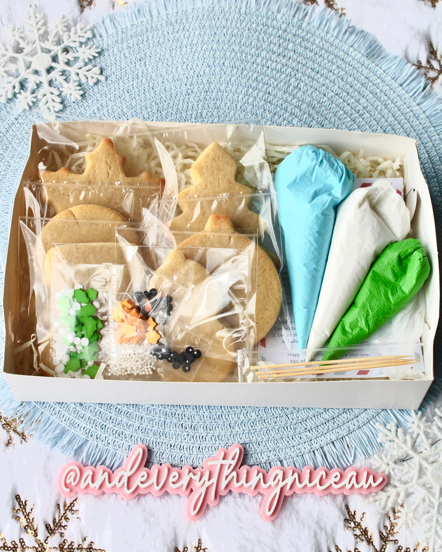 Open DIY Christmas cookie decorating kit showing six cookies, three icing bags in blue, green, and white, and a mix of sprinkles, displayed on a pale blue placemat over fluffy white fabric with gold snowflakes. Winter-themed, festive, and cozy styling.