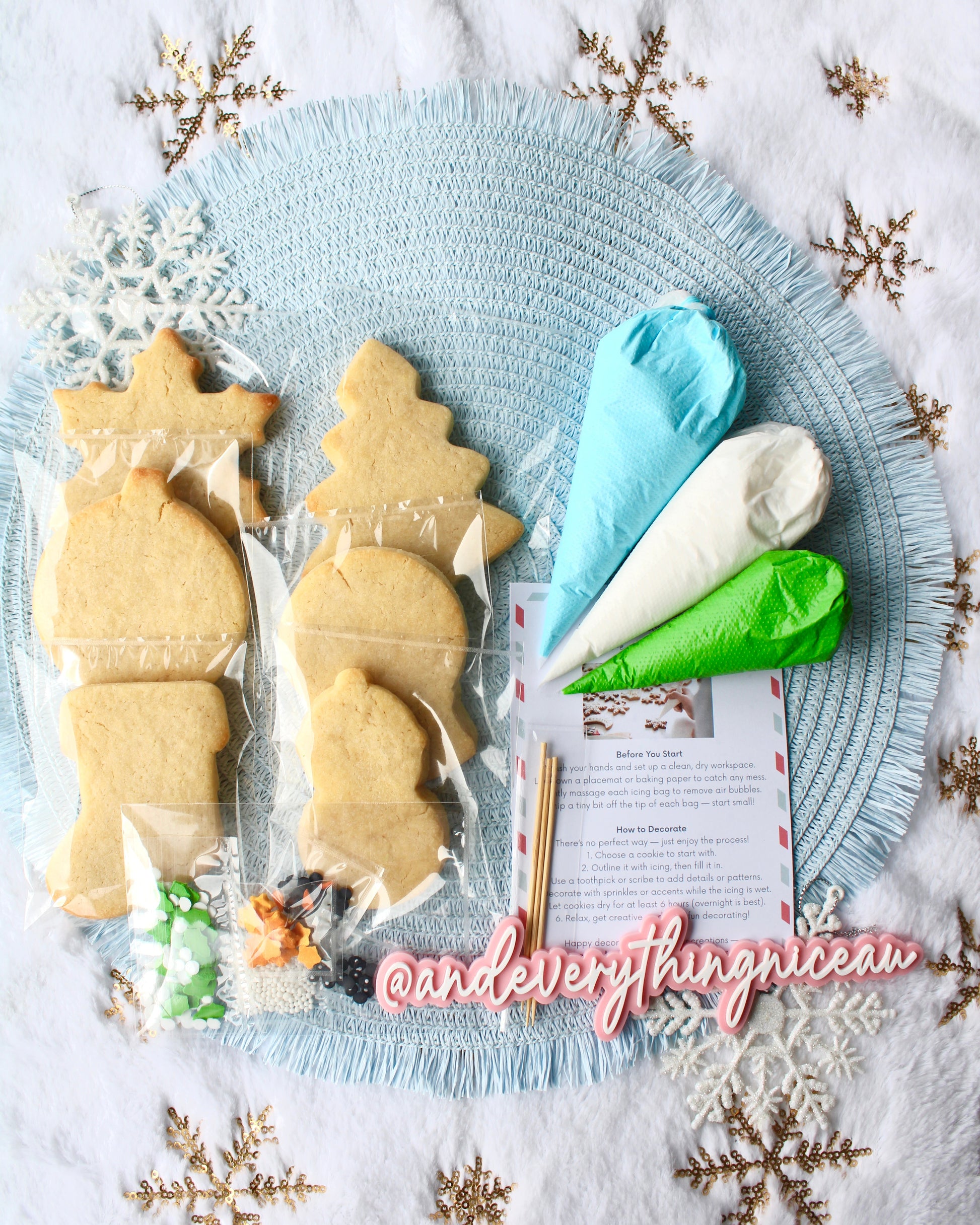 DIY Christmas cookie decorating kit with all items laid out, including six cookies, three icing bags in blue, green, and white, mixed sprinkles, decorating picks, and an instruction card, arranged on a pale blue placemat over fluffy white fabric with gold snowflakes.