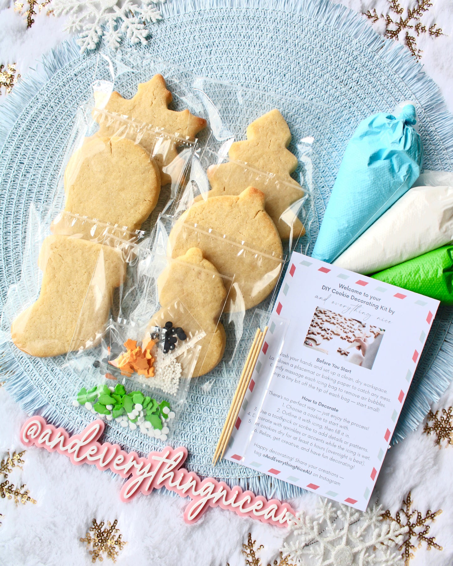 DIY Christmas cookie decorating kit with all items laid out, including six cookies, three icing bags in blue, green, and white, mixed sprinkles, decorating picks, and an instruction card, arranged on a pale blue placemat over fluffy white fabric with gold snowflakes.