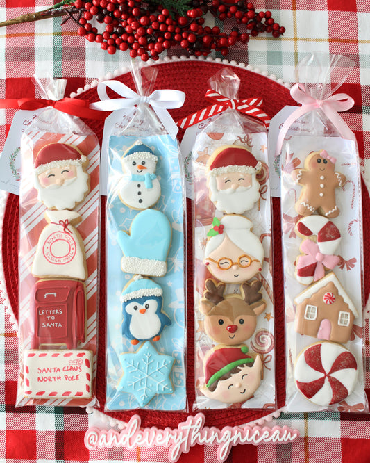 Four Christmas cookie packs, each in a different holiday-themed sleeve, tied with ribbons, styled on red and green tartan fabric