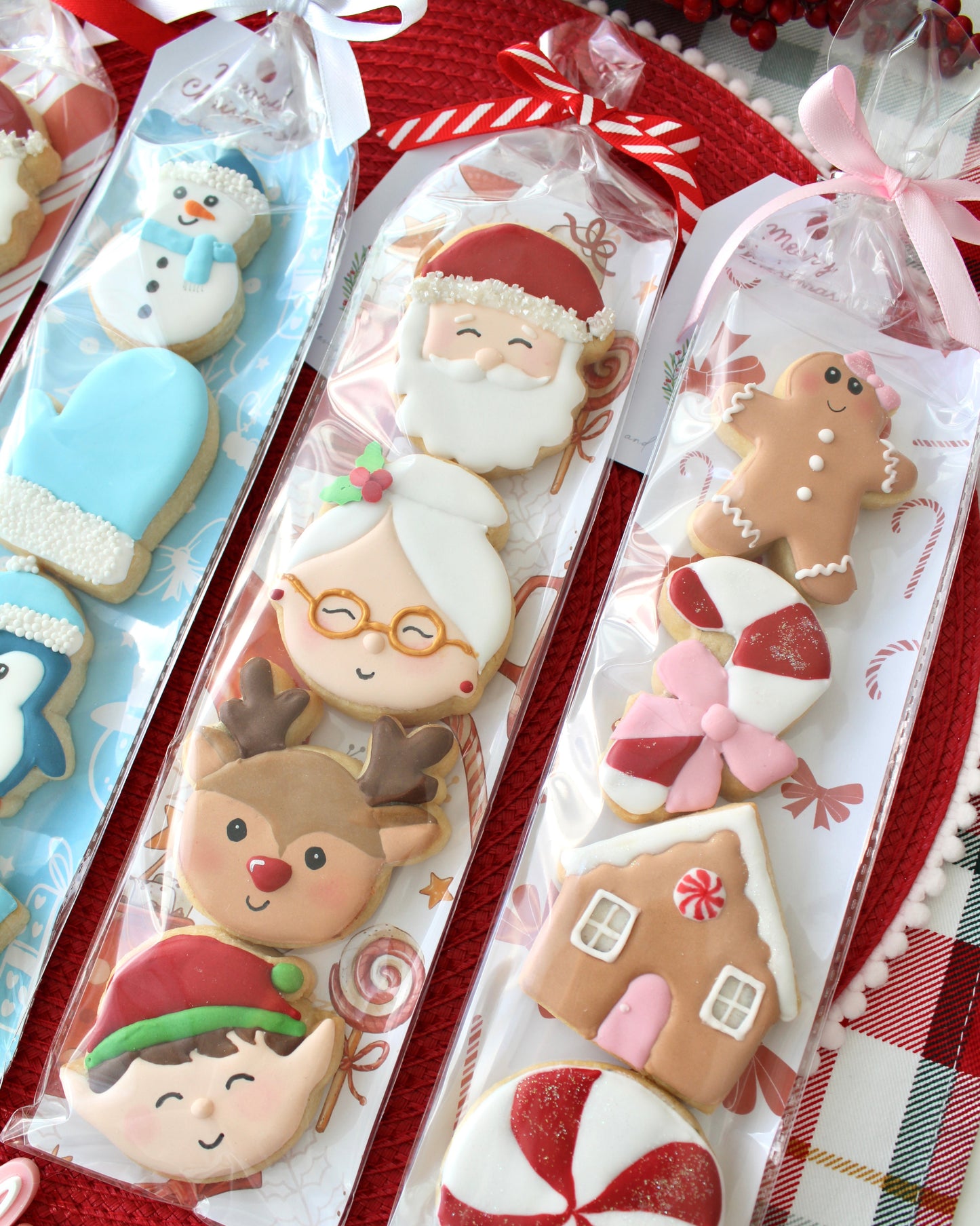 Close-up of two Christmas cookie packs: left pack includes Santa, Mrs Claus, reindeer, and elf face cookies tied with a red and white bow; right pack includes a gingerbread girl with bow, red and white candy cane with pink bow, gingerbread house with pink door, and red and white peppermint swirl cookie tied with a pink bow