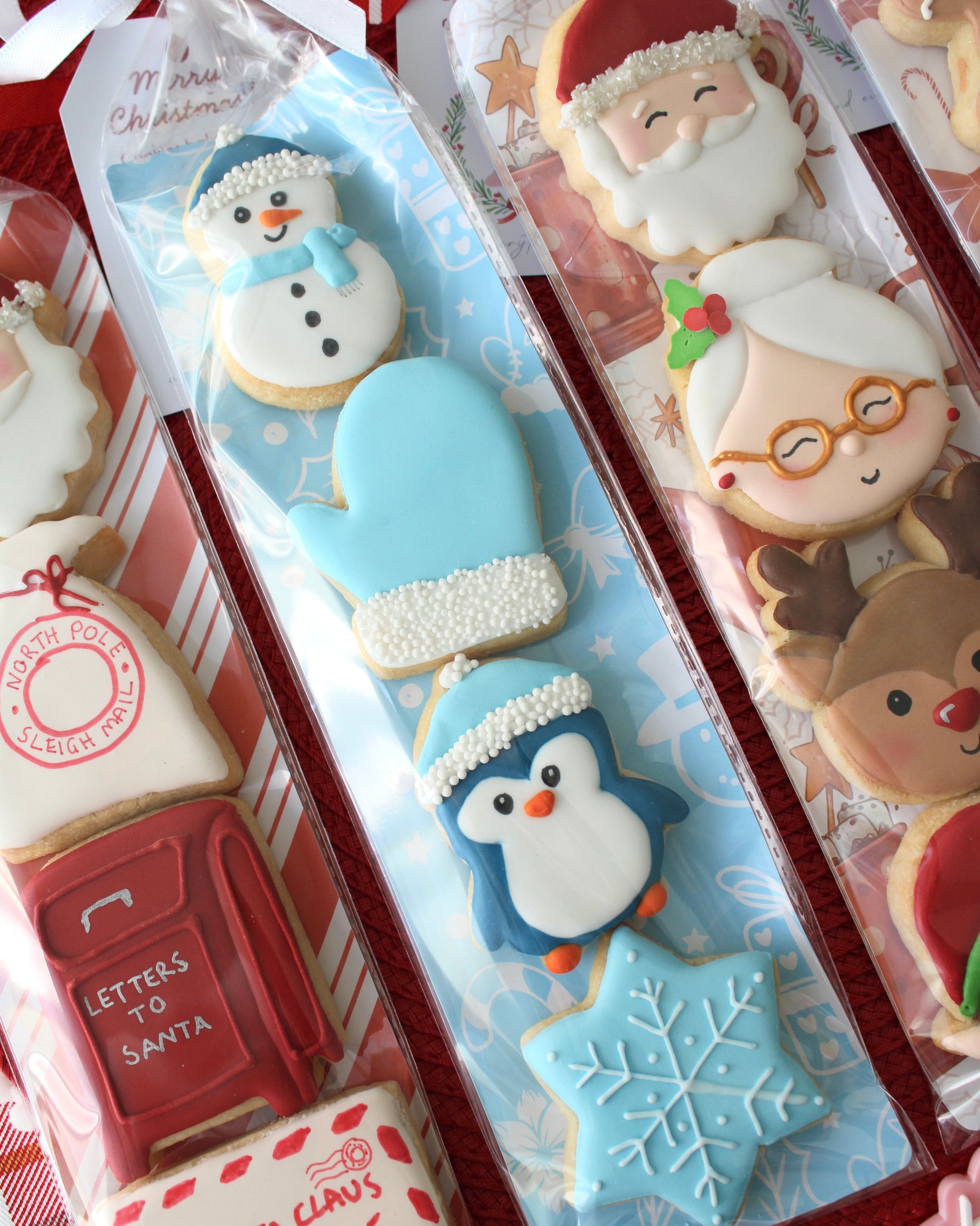 Close-up of a winter-themed cookie pack, including a snowman with pale blue beanie, pale blue mitten with white pearl cuff, dark blue penguin with pale blue beanie, and pale blue snowflake cookie, tied with a white bow