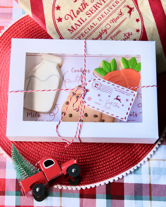 Closed white gift box tied with red and white twine and vintage gift tag, containing three large Christmas cookies from Santa’s Treat Box: a bottle of milk, a stack of cookies, and a bunch of carrots. The box sits on a red placemat over red and green tartan fabric, with a vintage mail-themed stocking in the top right corner and a red truck carrying a Christmas tree in the bottom left corner.