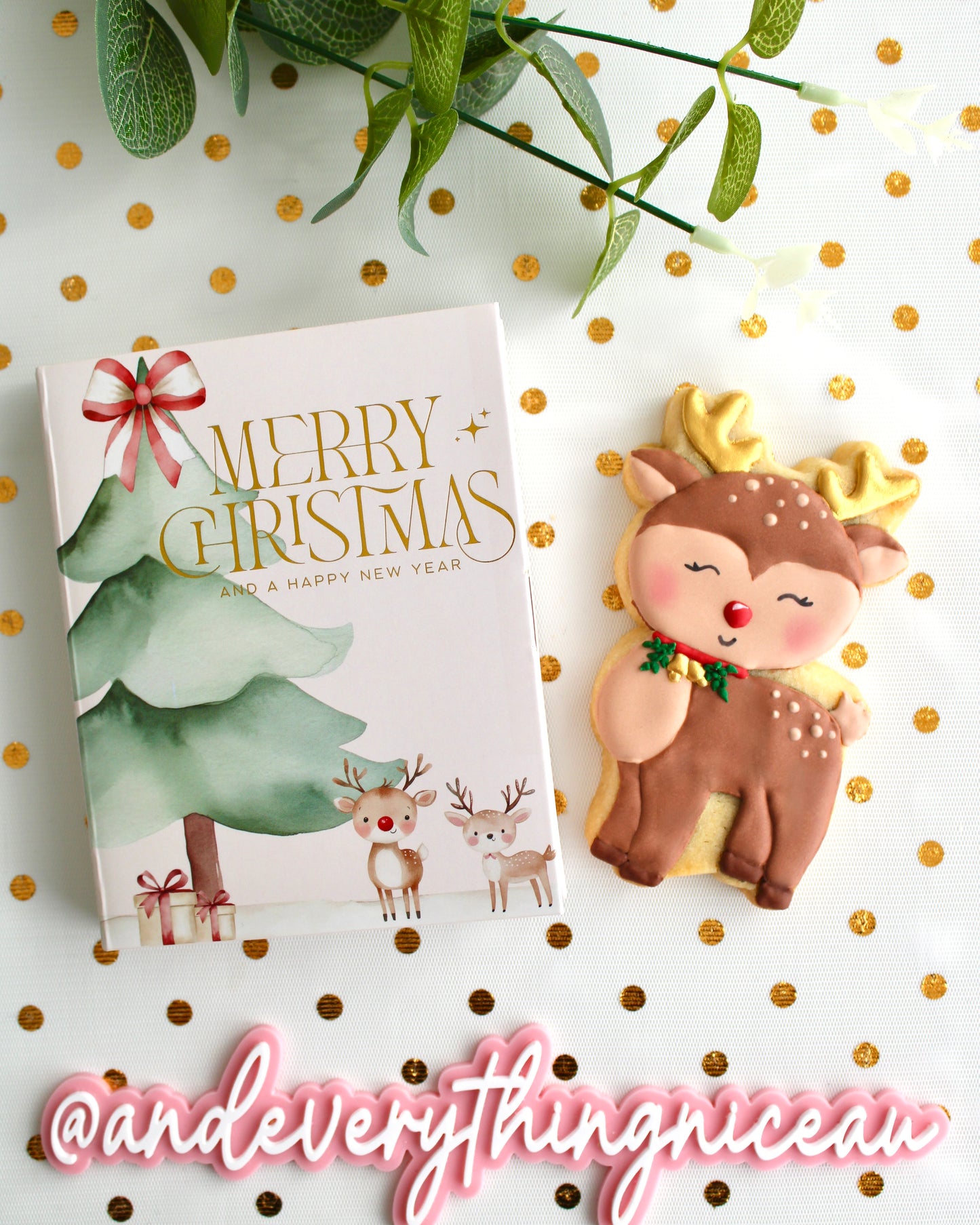 A festive Christmas cookie card box decorated with watercolour trees and reindeer sits on the left, and a brown and tan reindeer cookie with gold accents sits on the right. A pink and white “@andeverythingnice” physical watermark plaque is placed underneath. Everything is styled on white sheer fabric with gold dots.