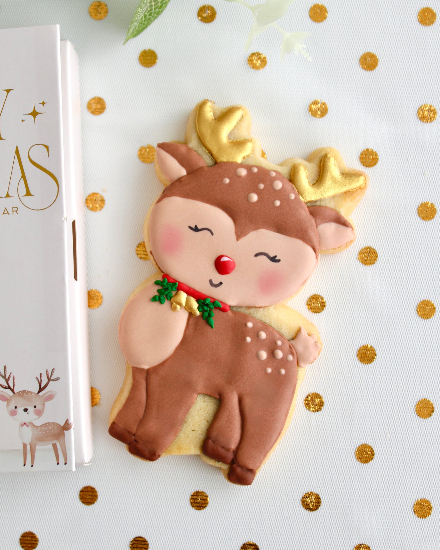 A brown and tan reindeer cookie with gold antlers, gold bells on a red collar with greenery, and a red nose, styled on sheer white fabric with gold dots.
