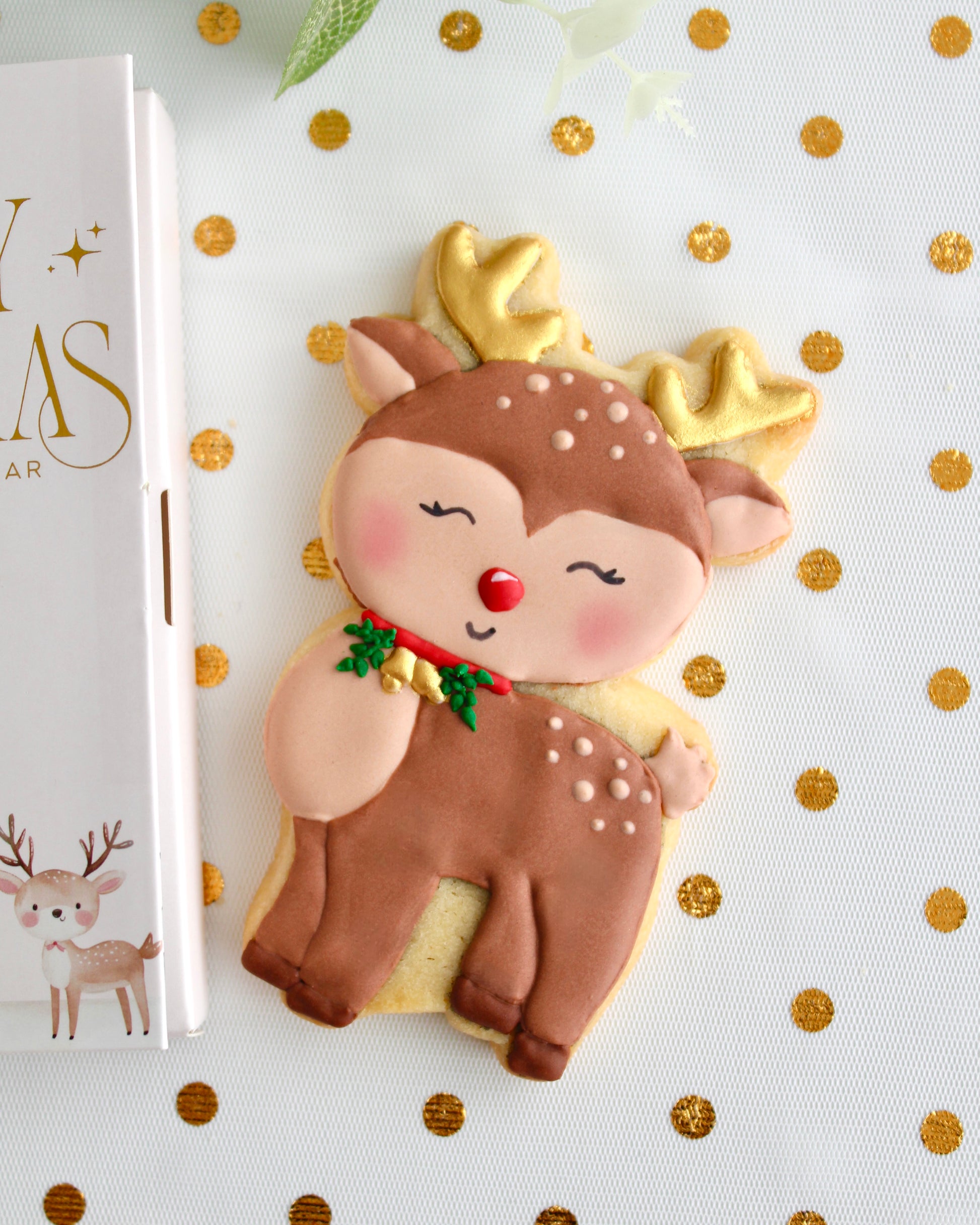 A brown and tan reindeer cookie with gold antlers, gold bells on a red collar with greenery, and a red nose, styled on sheer white fabric with gold dots.