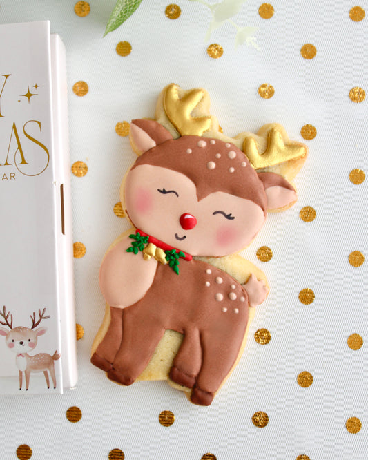 A brown and tan reindeer cookie with gold antlers, gold bells on a red collar with greenery, and a red nose, styled on sheer white fabric with gold dots.