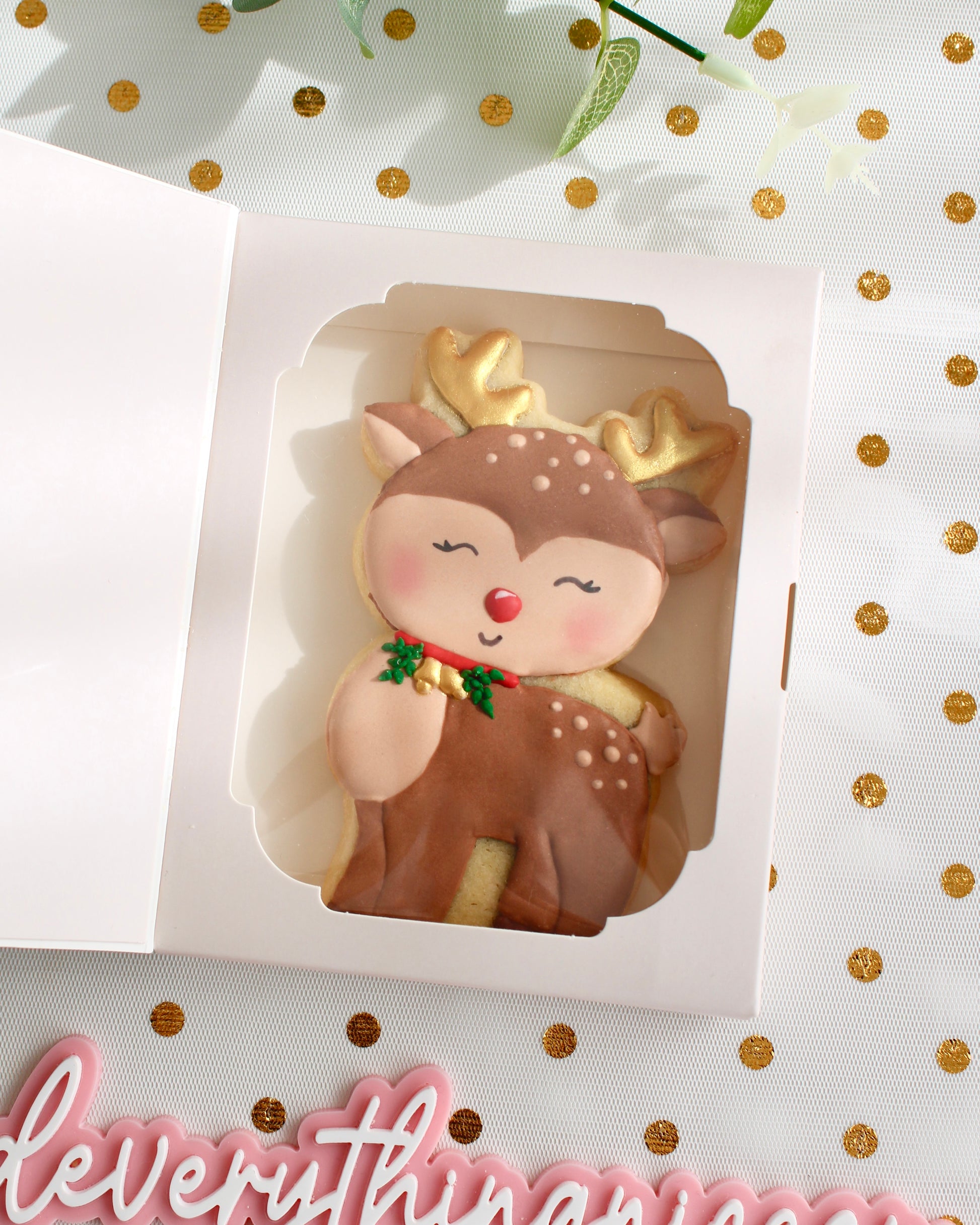 A brown and tan reindeer cookie with gold antlers, gold bells on a red collar with greenery, and a red nose, displayed inside a white cookie gift card box with a clear window, styled on sheer white fabric with gold dots.