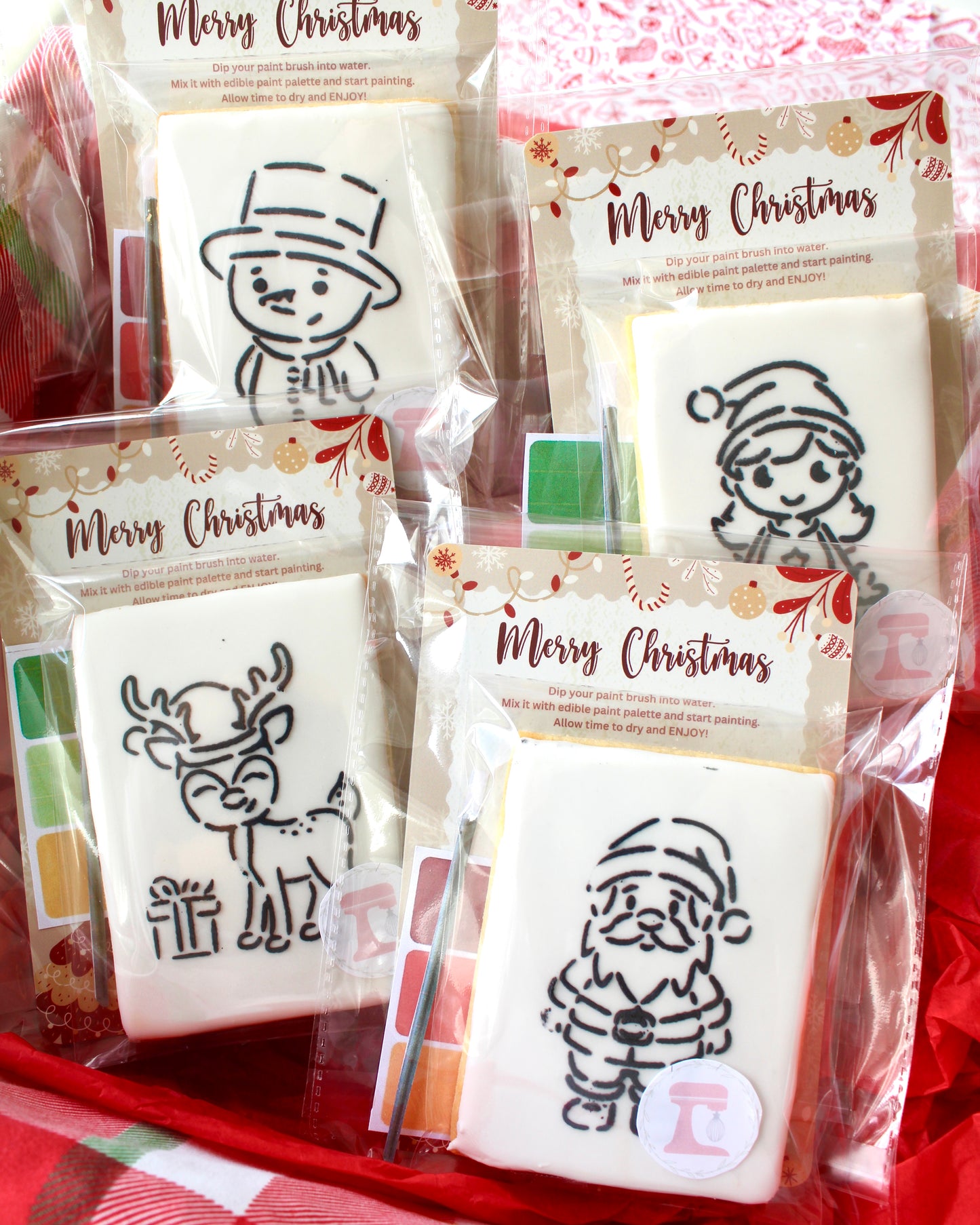 Four Christmas paint-your-own cookies featuring a snowman with a top hat, a Christmas girl holding presents, a reindeer with a gift, and Santa Claus. Each cookie is individually sealed with a festive backing card that says “Merry Christmas,” displayed on red fabrics with Christmas props.