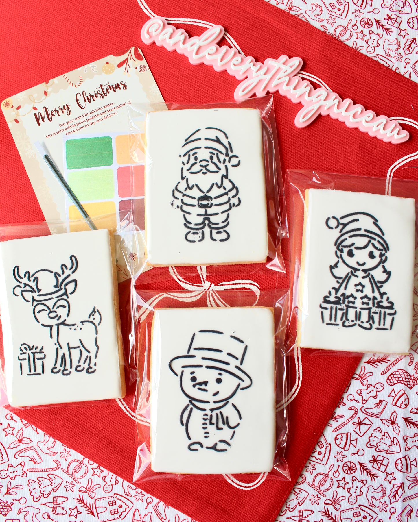 Four Christmas paint-your-own cookies featuring a snowman with a top hat, a Christmas girl holding presents, a reindeer with a gift, and Santa Claus. Each cookie is individually sealed, with a festive “Merry Christmas” backing card in the background, a paintbrush, and a mini paint palette in Christmas colours, displayed on red fabrics.