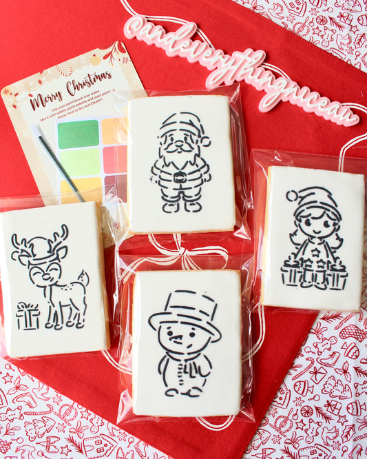 Four Christmas paint-your-own cookies featuring a snowman with a top hat, a Christmas girl holding presents, a reindeer with a gift, and Santa Claus. Each cookie is individually sealed, with a festive “Merry Christmas” backing card in the background, a paintbrush, and a mini paint palette in Christmas colours, displayed on red fabrics.