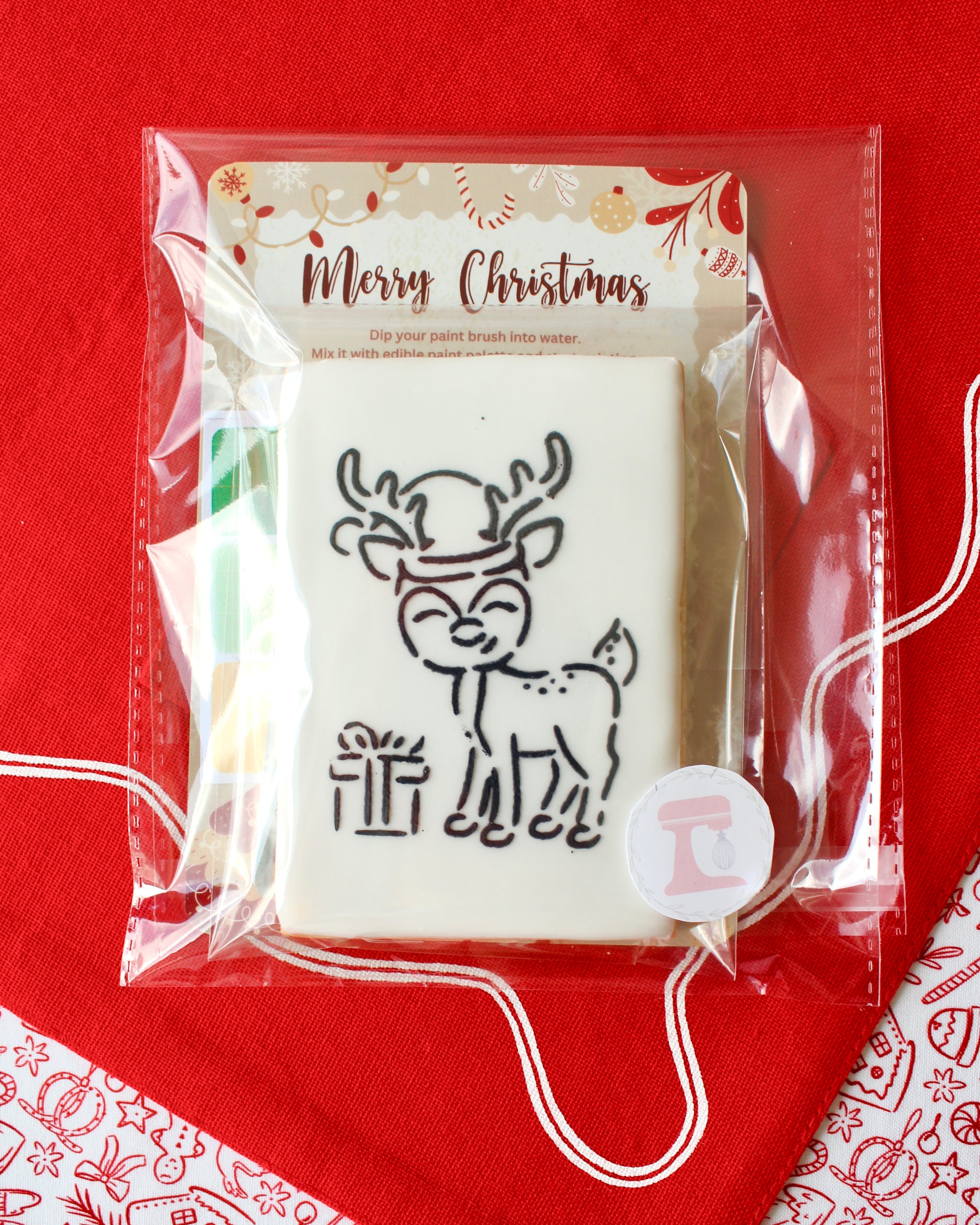 Close-up of a Christmas paint-your-own cookie featuring a reindeer with a present, individually sealed with a festive “Merry Christmas” backing card, a paintbrush, and a mini paint palette in Christmas colours, all included in the pack and displayed on red Christmas fabrics.