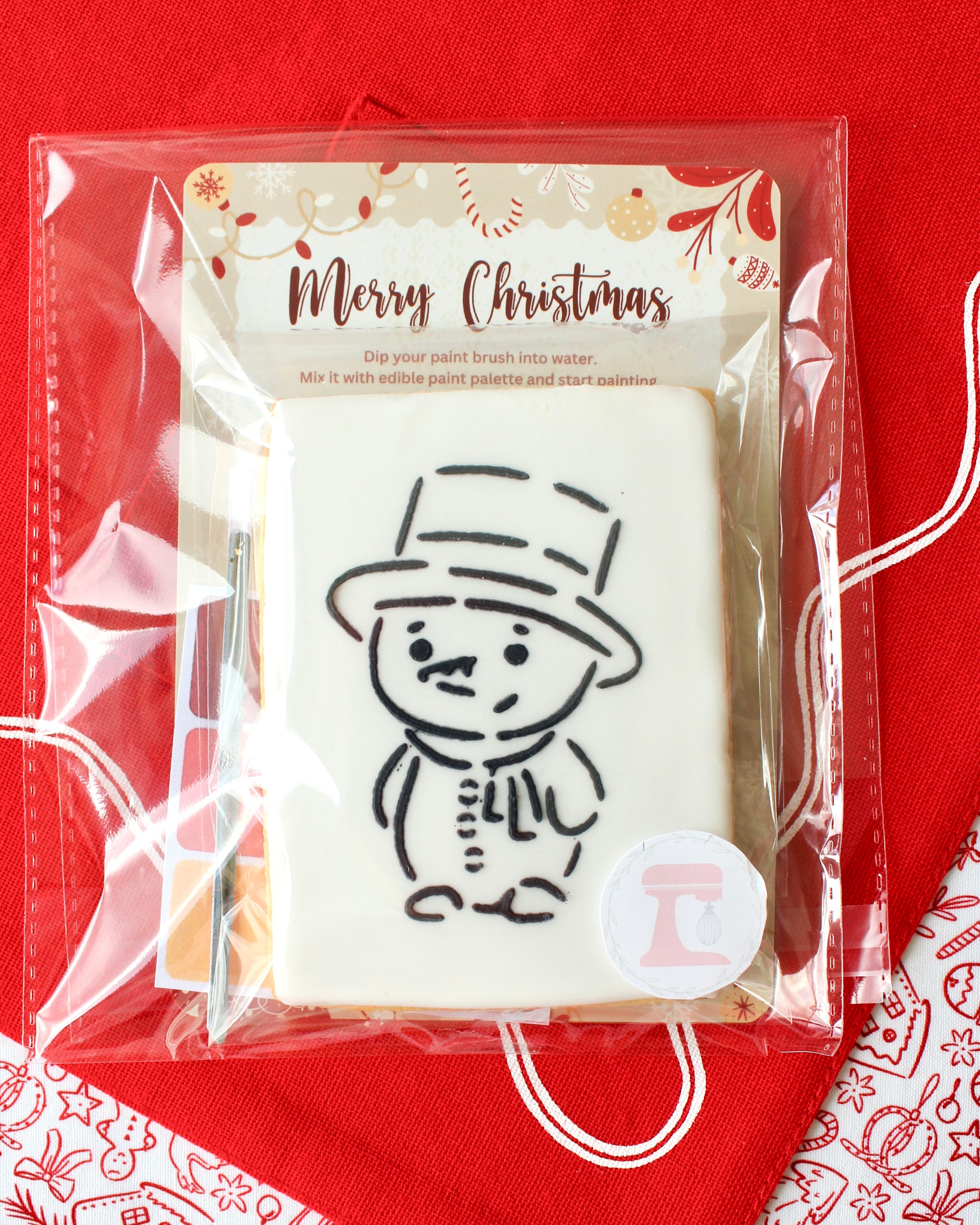 Close-up of a Christmas paint-your-own cookie featuring a snowman with a top hat, individually sealed with a festive “Merry Christmas” backing card, a paintbrush, and a mini paint palette in Christmas colours, all included in the pack and displayed on red Christmas fabrics.