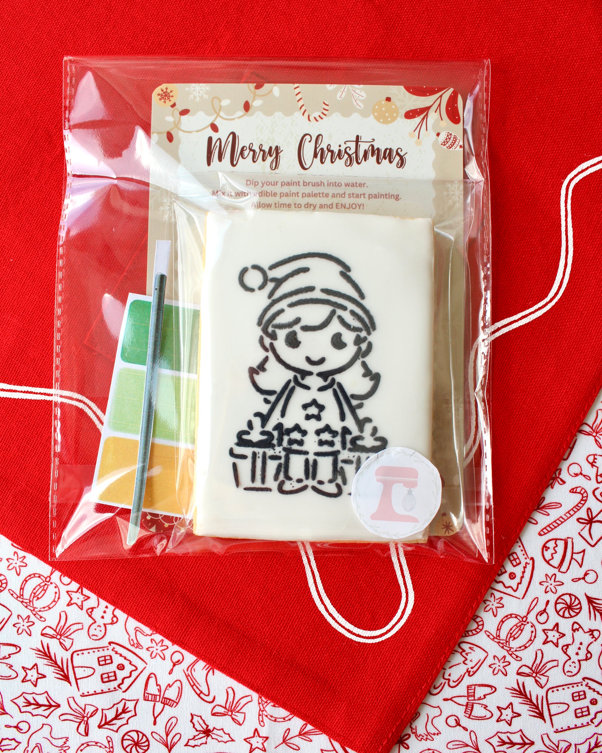 Close-up of a Christmas paint-your-own cookie featuring a Christmas girl holding two presents, individually sealed with a festive “Merry Christmas” backing card, a paintbrush, and a mini paint palette in Christmas colours, all included in the pack and displayed on red festive fabrics.