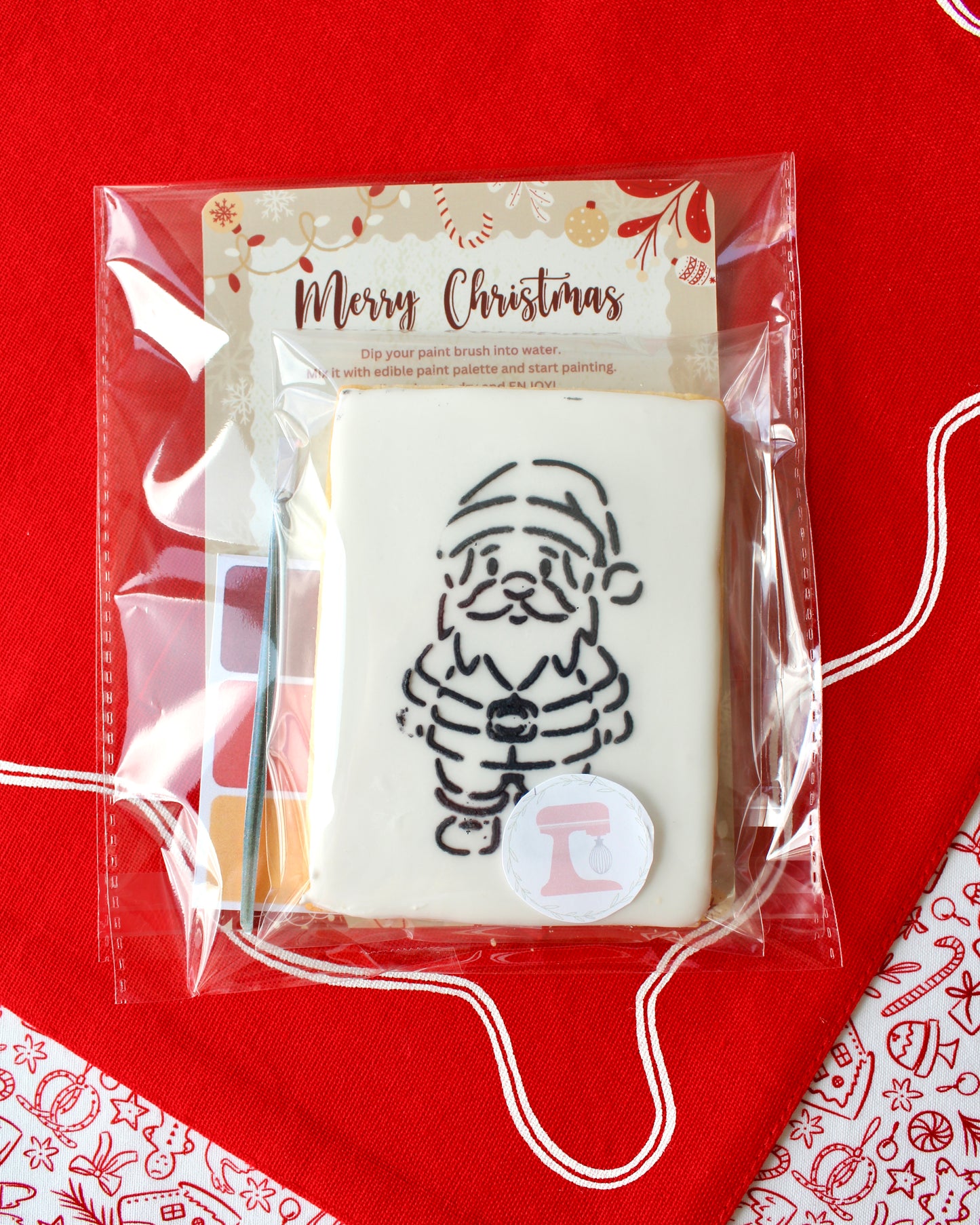 Close-up of a Christmas paint-your-own cookie featuring Santa Claus, individually sealed with a festive “Merry Christmas” backing card, a paintbrush, and a mini paint palette in Christmas colours, all included in the pack and displayed on red festive fabrics.