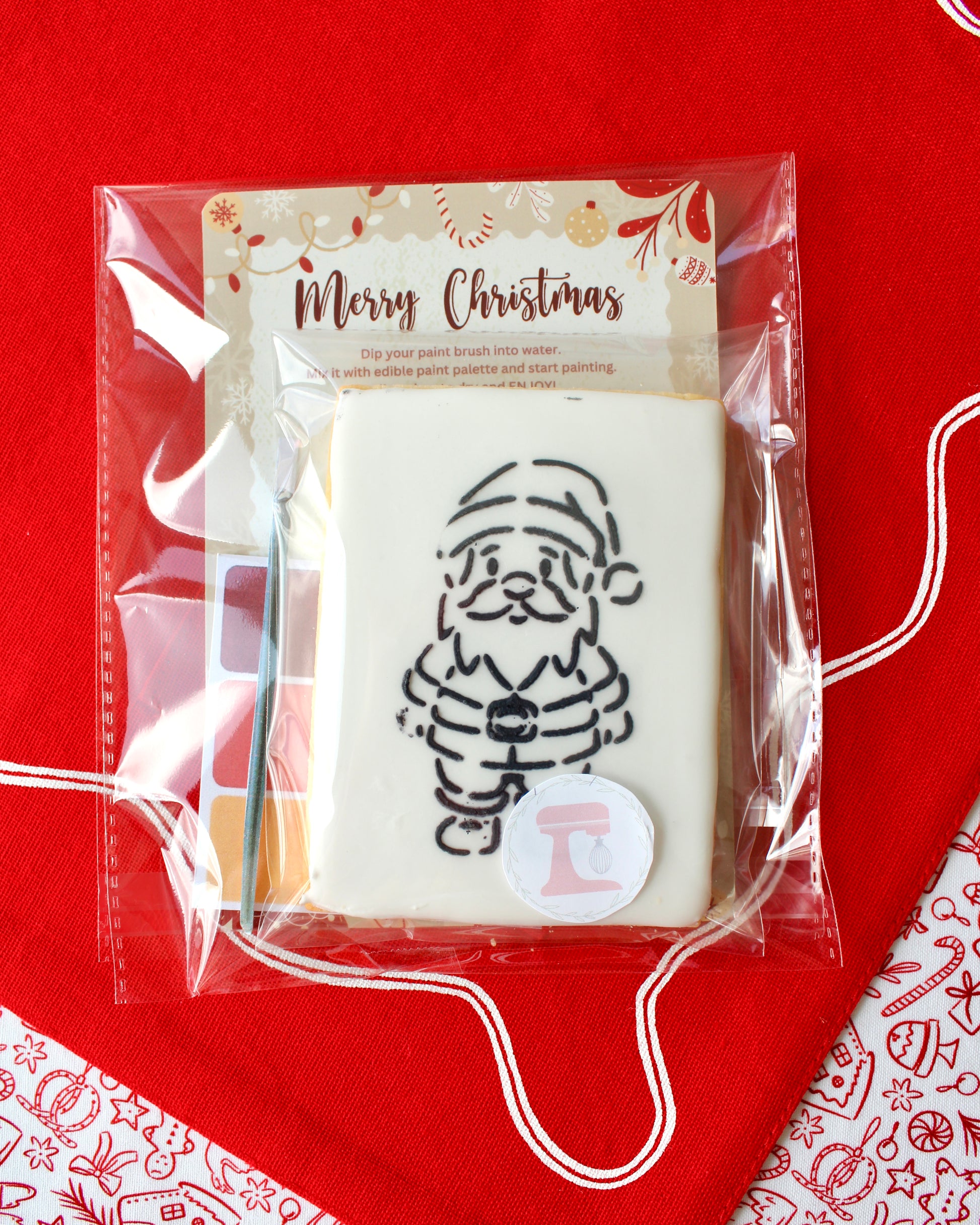 Close-up of a Christmas paint-your-own cookie featuring Santa Claus, individually sealed with a festive “Merry Christmas” backing card, a paintbrush, and a mini paint palette in Christmas colours, all included in the pack and displayed on red festive fabrics.