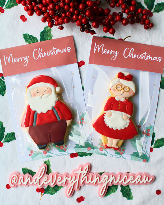 Two Christmas cookie packs displayed side by side. One features a Santa cookie wearing a red long-sleeve top, maroon pants, grey suspenders, black shoes, his classic white beard, and a red and white Santa hat. The other features a Mrs Claus cookie wearing a tea-length red dress with a white collar and gold buttons, her white hair in a bun with a red bow. Both cookies are packaged with Christmas backing cards that read “Merry Christmas” at the top.