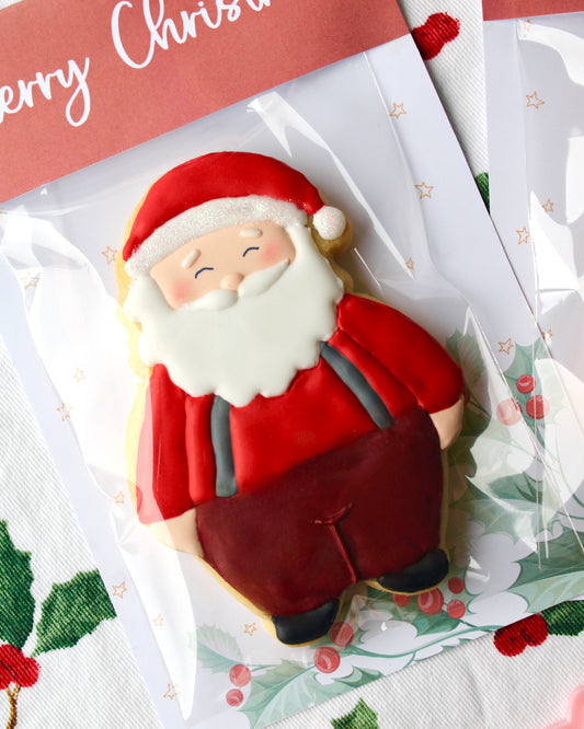 Close-up of a Santa Claus cookie wearing a red long-sleeve top, maroon pants, grey suspenders, black shoes, a classic white beard, and a red and white Santa hat.
