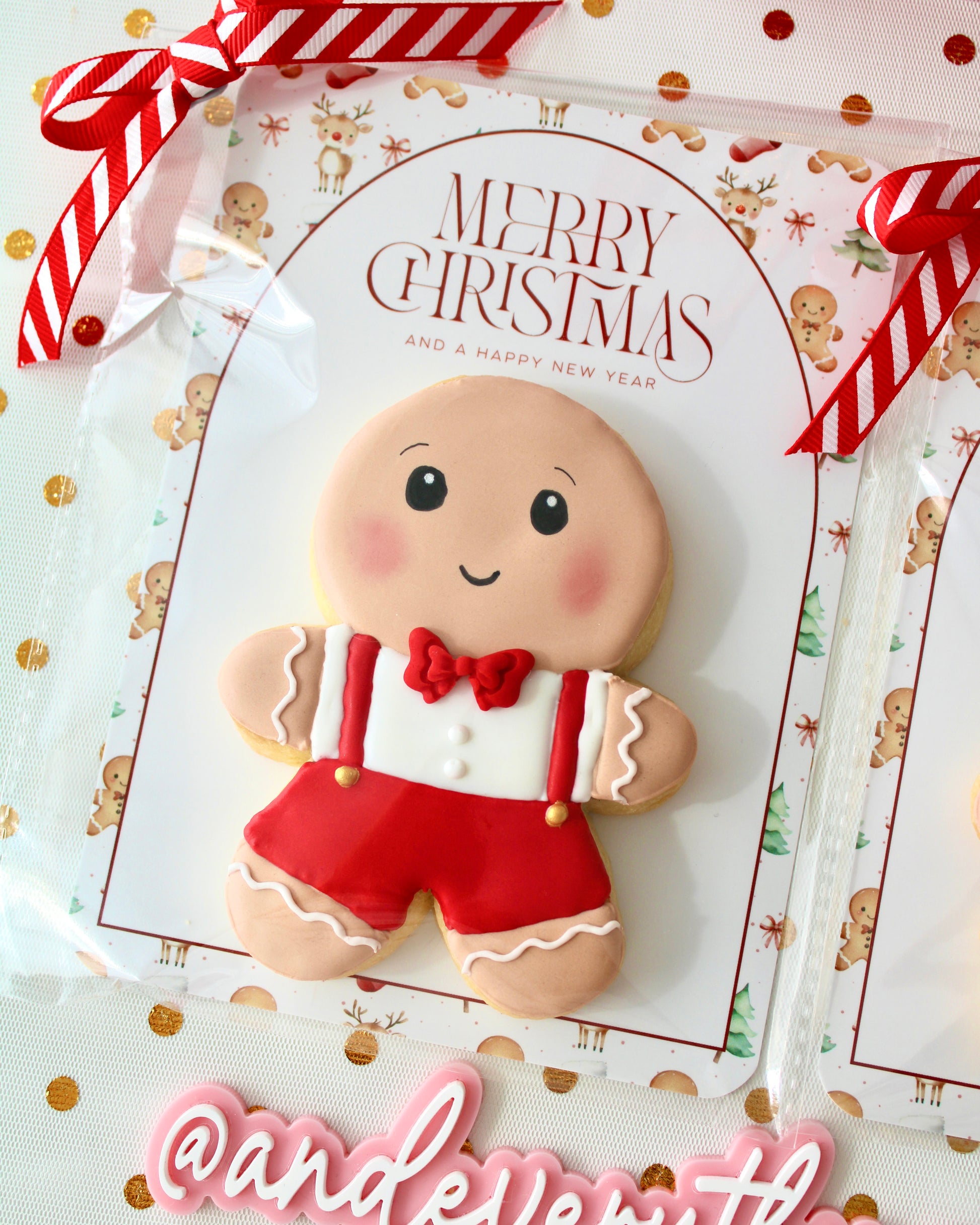 Close-up of a gingerbread boy cookie featuring a red bow tie, white t-shirt, and red suspenders with gold buttons. The cookie is individually wrapped in a clear bag with a festive backing card and tied with a red and white ribbon, styled on sheer white fabric decorated with gold metallic dots.