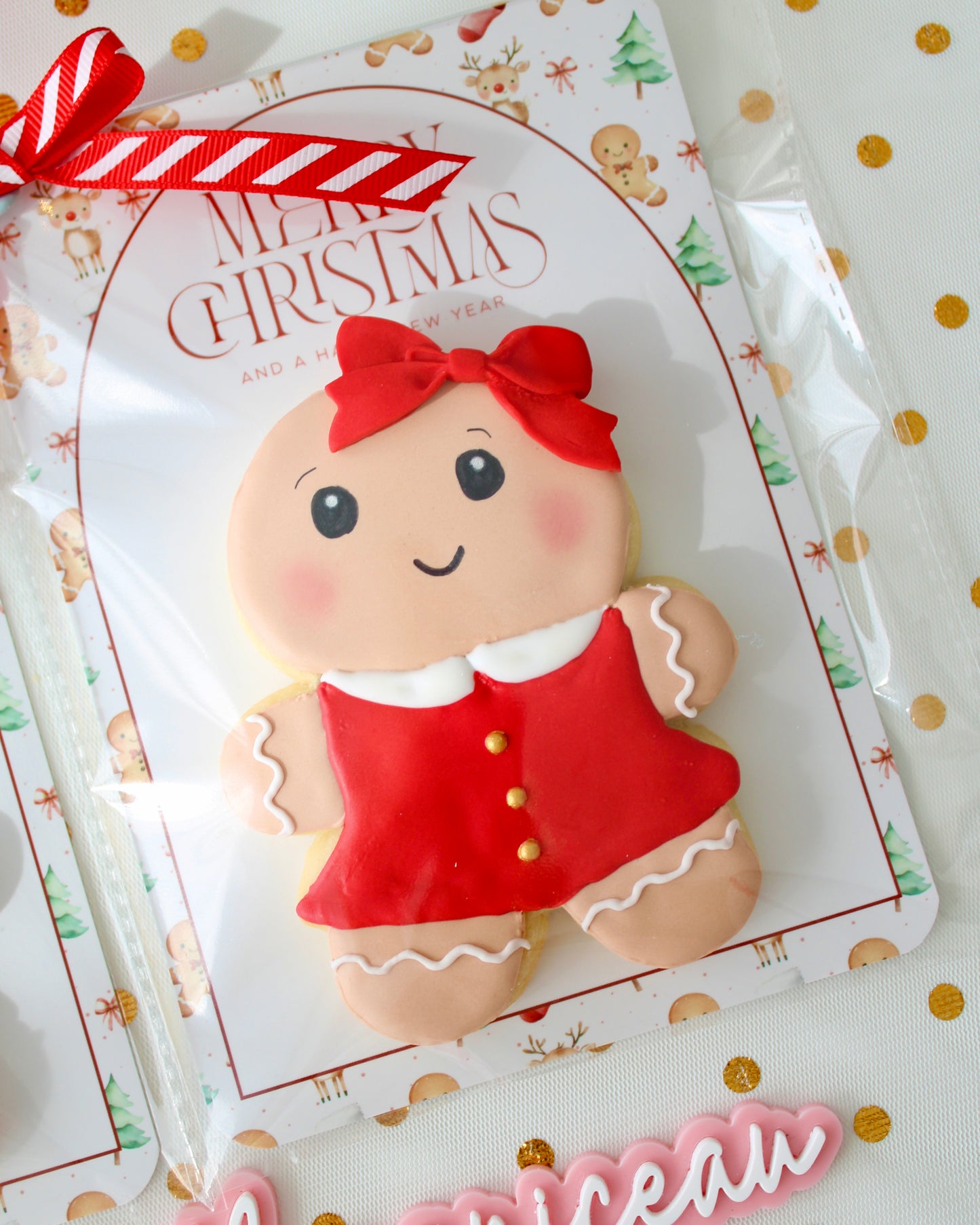 Close-up of a gingerbread girl cookie wearing a red dress with a white collar, gold buttons, and a red bow in her hair. The cookie is individually wrapped in a clear bag with a festive backing card and tied with a red and white ribbon, styled on sheer white fabric decorated with gold metallic dots.