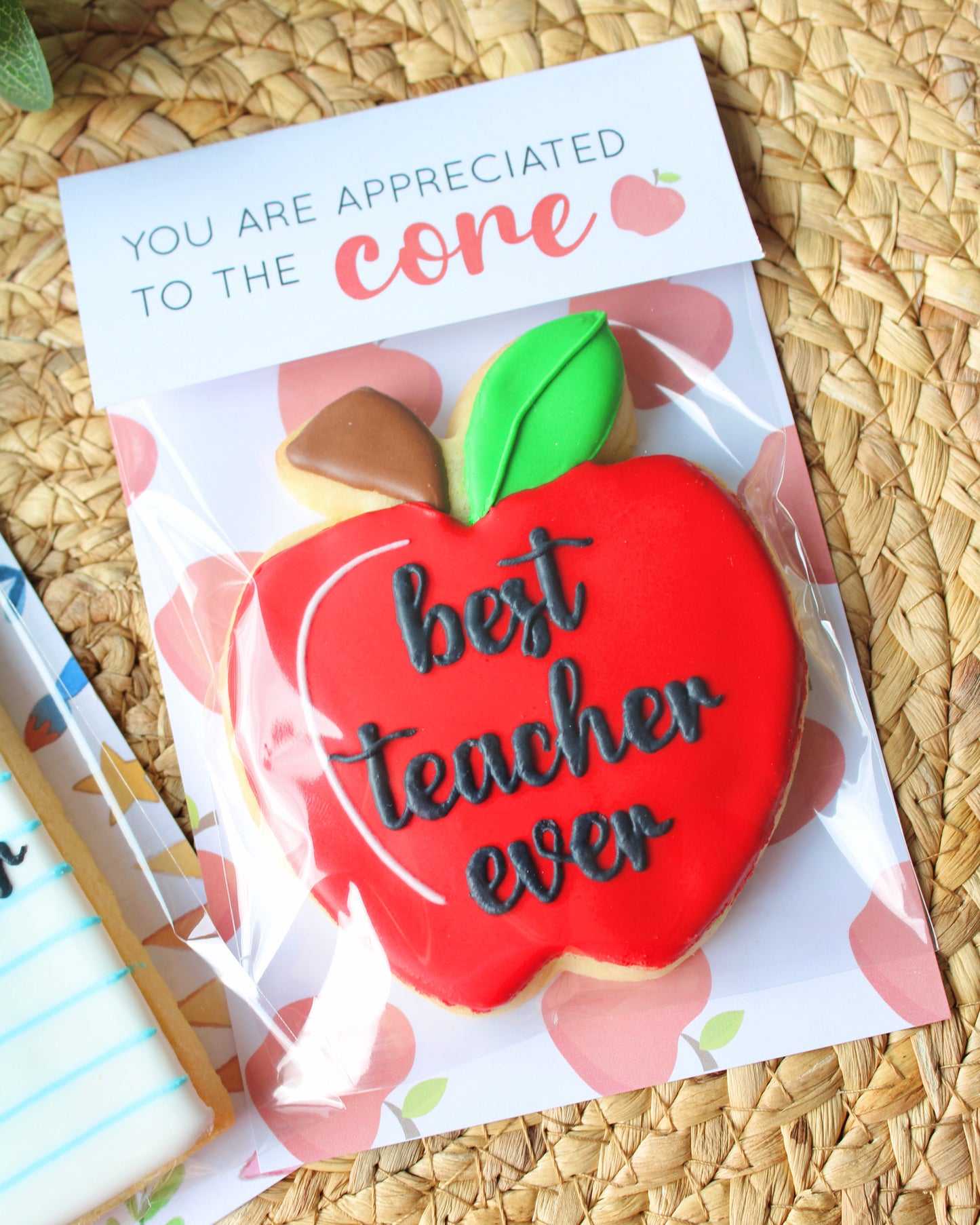 Close-up of a teacher appreciation cookie styled on a natural hyacinth placemat with eucalyptus sprigs. The cookie is shaped like a red apple with a green leaf and the words “best teacher ever” in black script icing. The apple cookie’s topper reads “You are appreciated to the core.