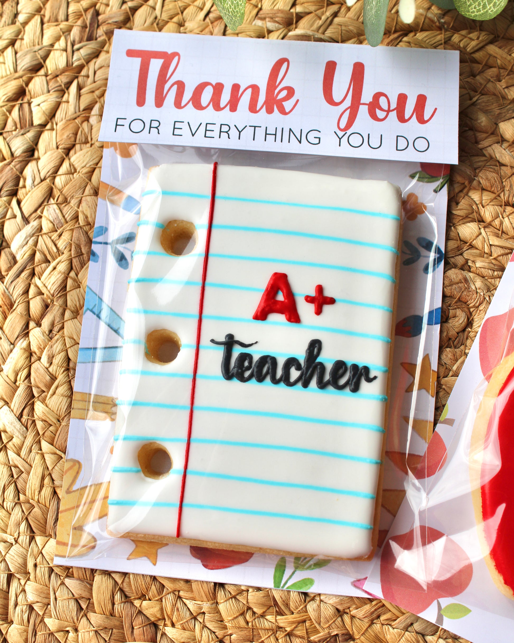 An individually packaged teacher appreciation cookie shaped like a white notepad with blue lines, featuring “A+” written in red and “teacher” in black script icing. The notepad cookie’s topper reads “Thank you for everything you do,” and it’s styled on a natural hyacinth placemat with eucalyptus sprigs.