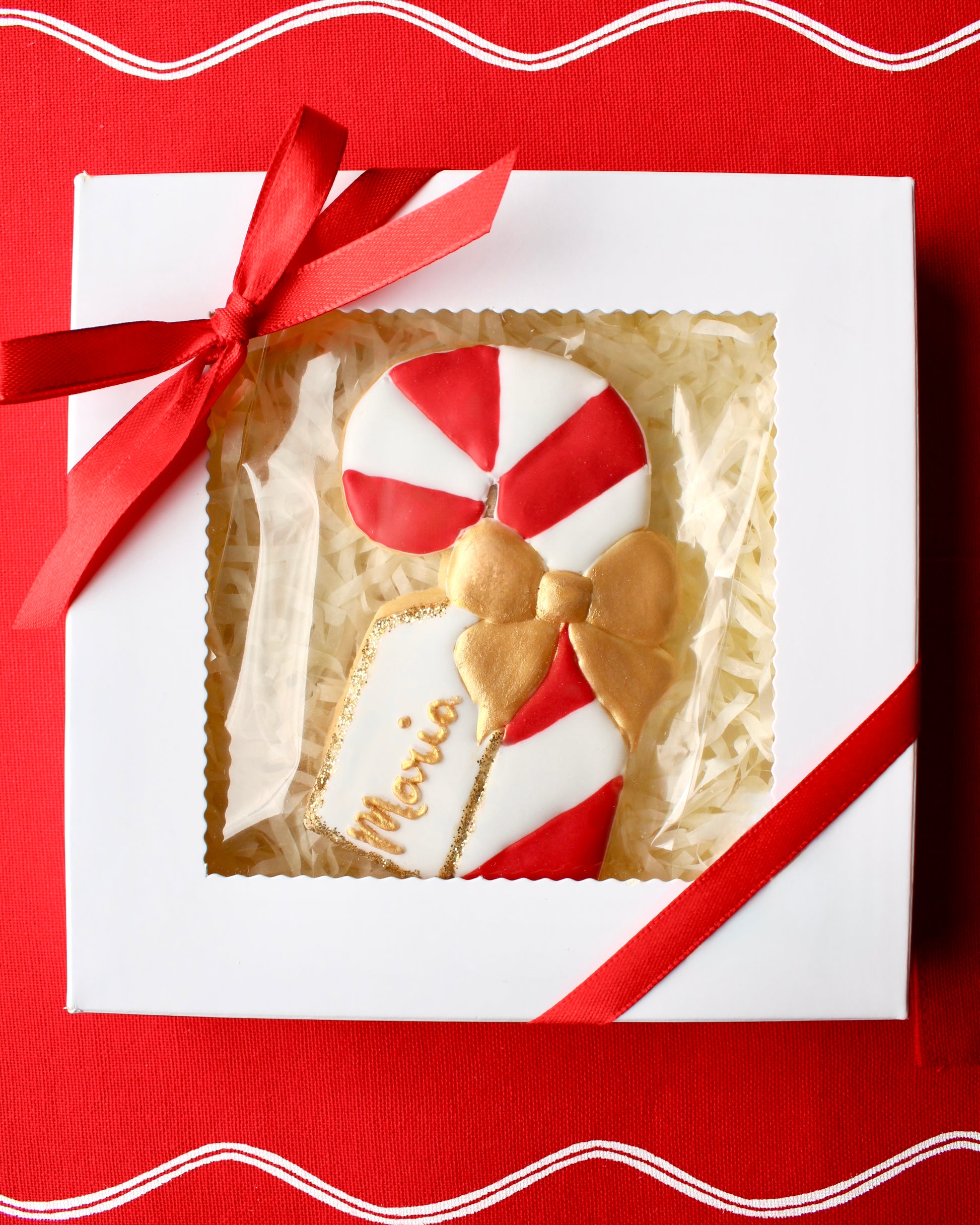 Red and white candy cane cookie decorated with a gold icing bow and a white edible name tag that says "Maria" in gold lettering with edible gold glitter edges. The cookie is placed inside a white box with a clear window, tied with red ribbons at the top left and bottom right corners, styled on red linen fabric.