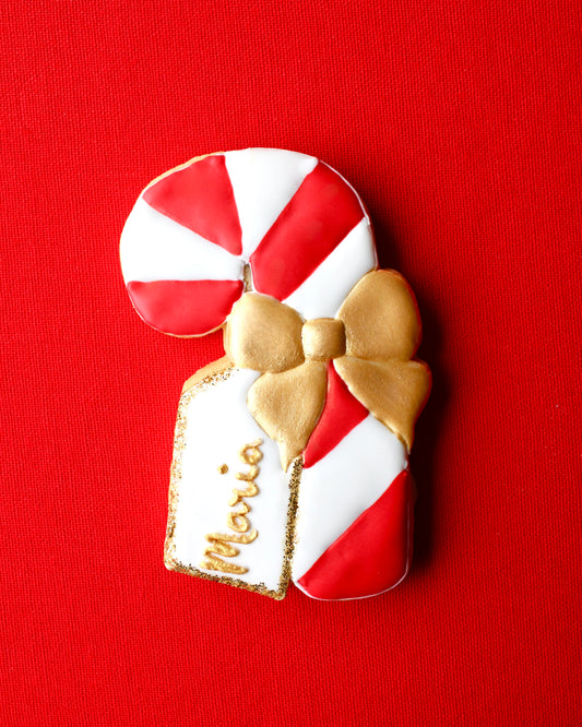 Close-up of a red and white candy cane cookie decorated with a gold icing bow and a white edible name tag that says "Maria" in gold lettering with edible gold glitter edges, styled on red linen fabric.