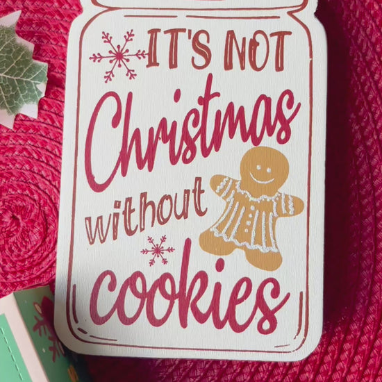 12 Days of Christmas cookie box featuring 12 mini Christmas-themed cookies arranged around the box, with a red cookie on top reading “Merry Christmas” in white cursive lettering. The cookies are decorated in festive red, green, white, and gold colours.