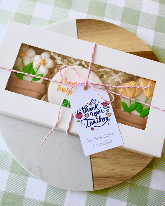 A teacher appreciation cookie box with three flower and garden-themed cookies inside, tied with pink and white twine. A gift card attached reads ‘Thank You Teacher’ with school-themed graphics. The box is styled on a marble and wood platter with green and white checkered fabric underneath.