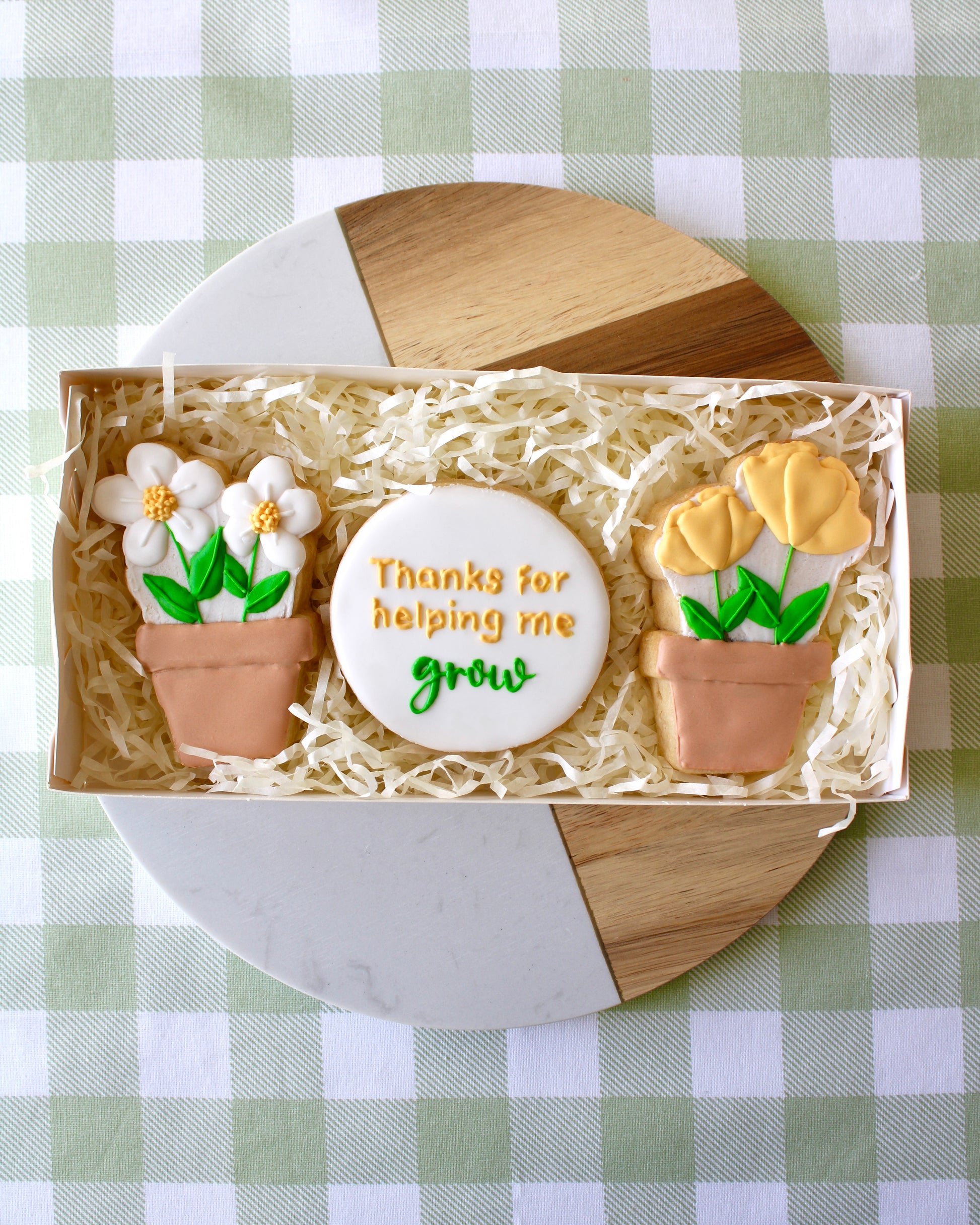 A teacher appreciation cookie box opened to show three cookies: a terracotta pot with white daisies, a round white cookie piped with ‘thank you for helping me grow’ in yellow and green royal icing, and a terracotta pot cookie with yellow flowers. The box is styled on a marble and wood platter with green and white checkered fabric underneath.