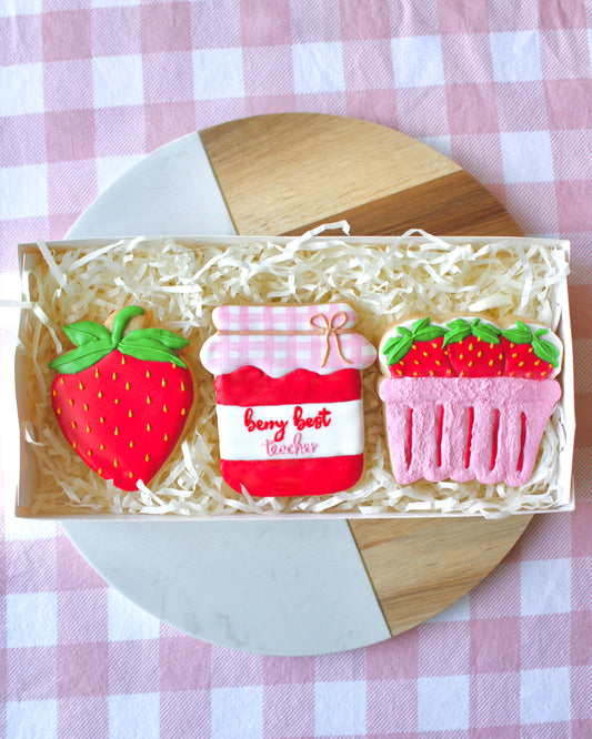 A teacher appreciation cookie box opened to show three cookies: a red strawberry with yellow seeds and green leaves, a strawberry jam jar cookie labeled ‘berry best teacher’ in red and pink royal icing with a white and pink checkered lid, and a pink strawberry punnet featuring three red strawberries. The box is styled on a marble and wood platter with pink and white checkered fabric underneath.