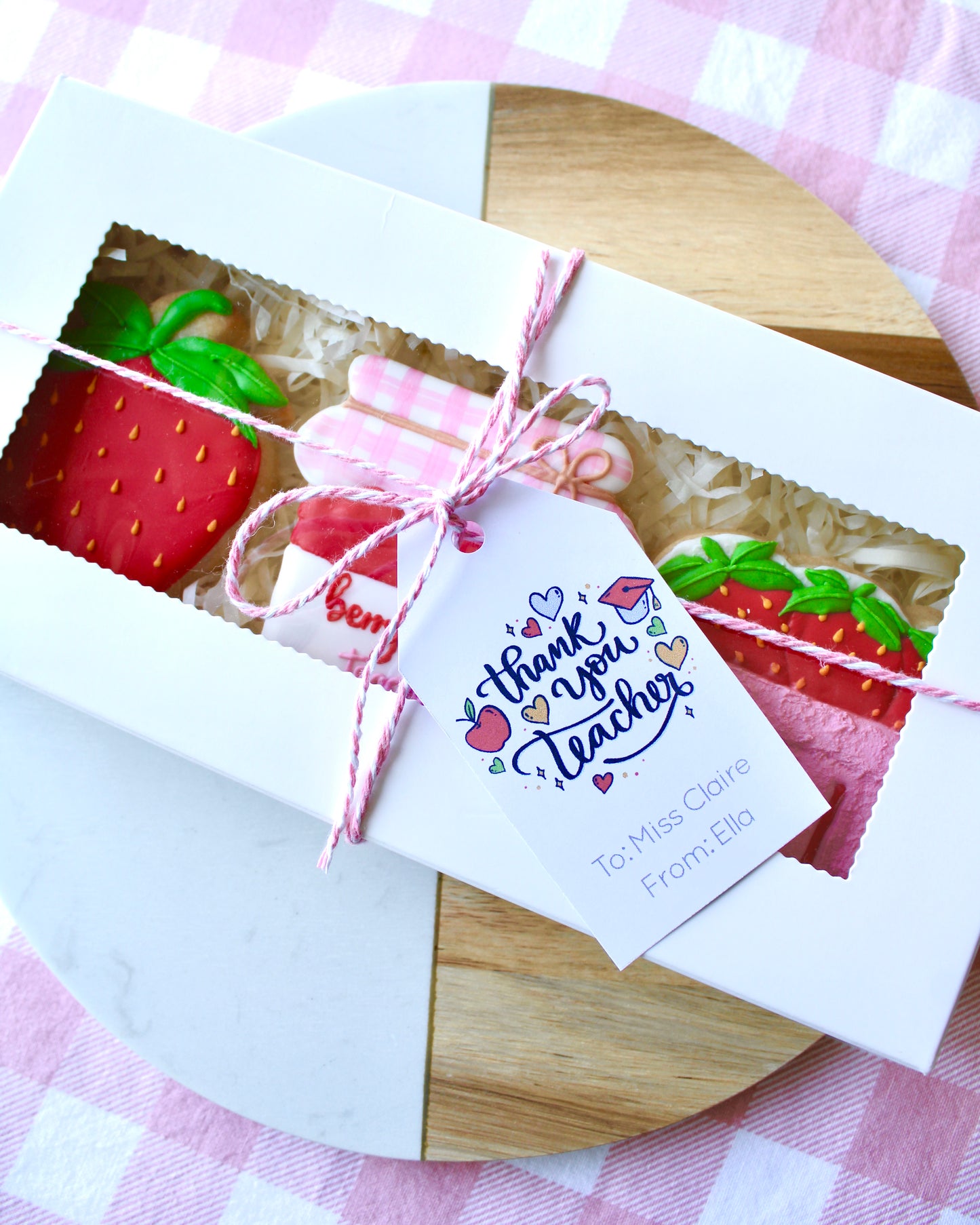 A teacher appreciation cookie box tied with pink and white twine, containing three strawberry-themed cookies. A gift card reading ‘Thank You Teacher’ with school-themed graphics is attached. The box is styled on a marble and wood platter with pink and white checkered fabric underneath.