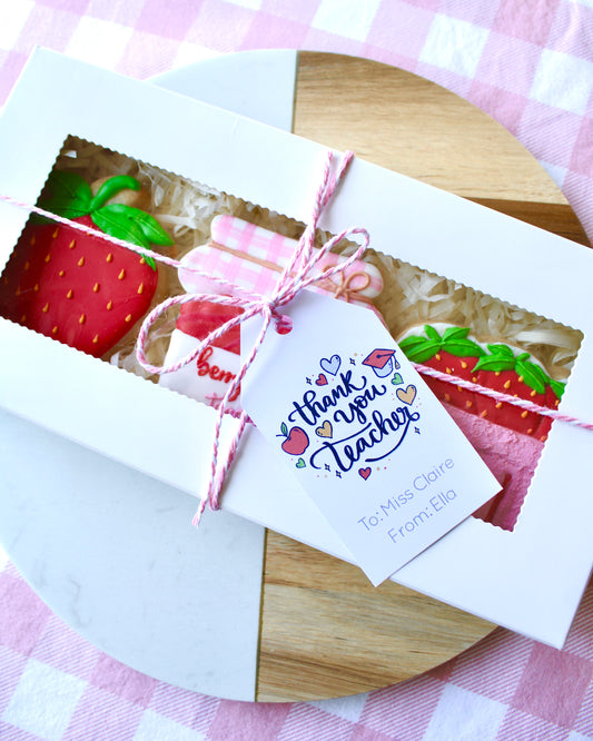 A teacher appreciation cookie box tied with pink and white twine, containing three strawberry-themed cookies. A gift card reading ‘Thank You Teacher’ with school-themed graphics is attached. The box is styled on a marble and wood platter with pink and white checkered fabric underneath.